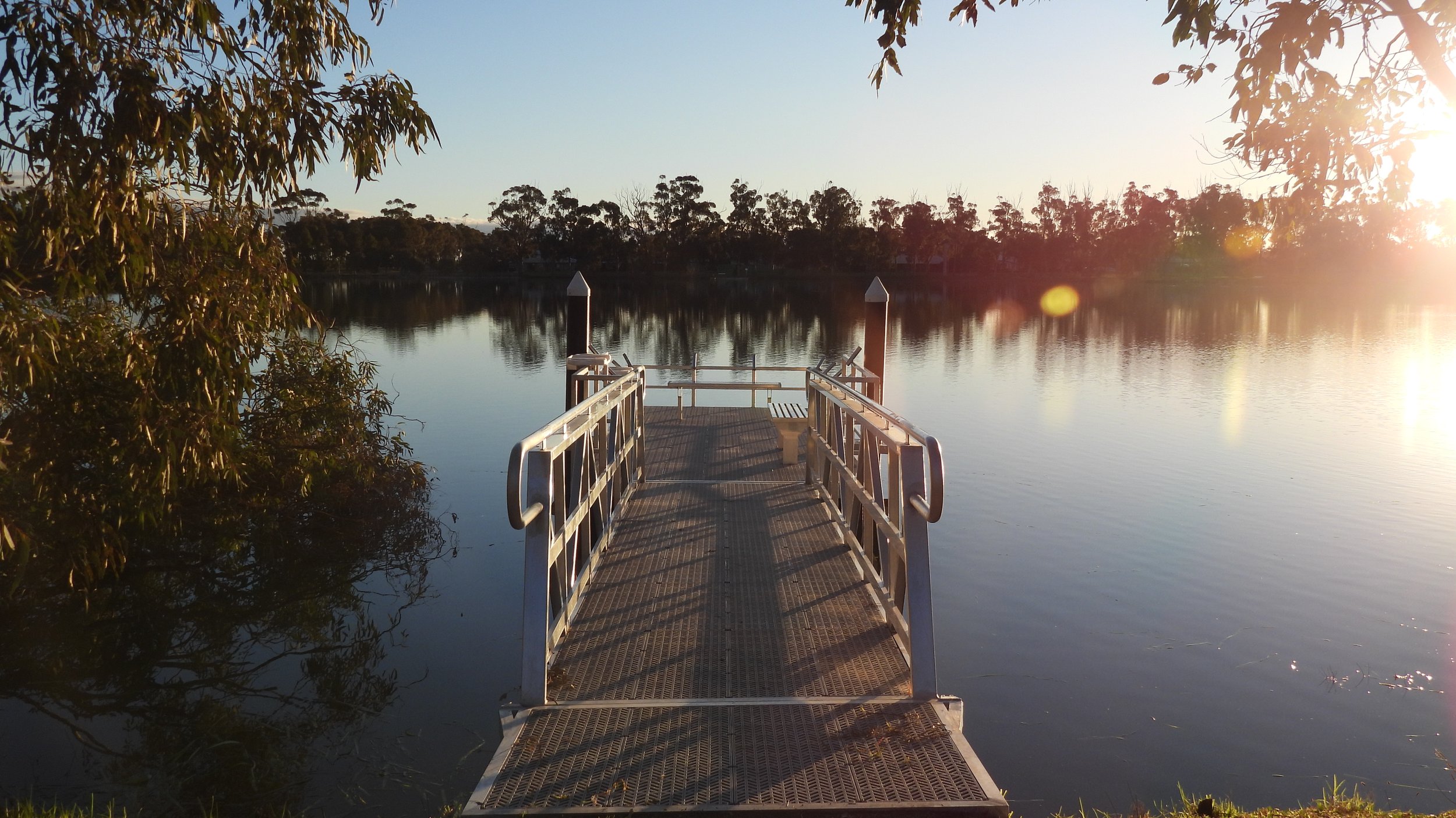 A metal dock extends out over calm water at sunset, with trees in the background and sun flare on the right side of the image.