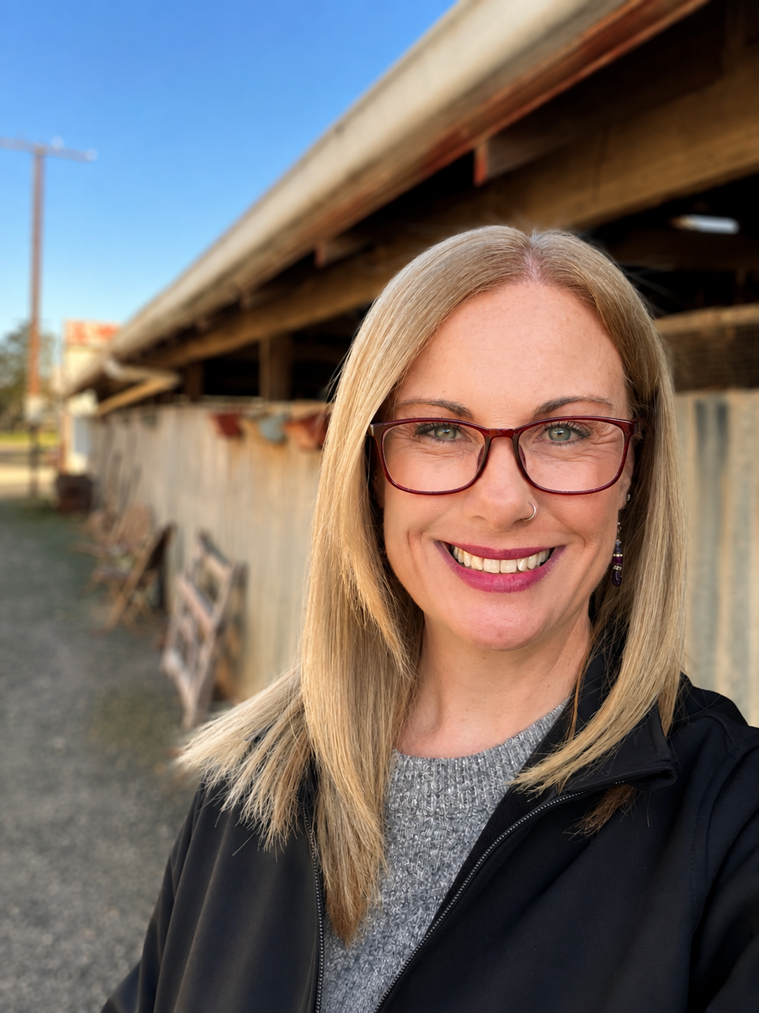 A smiling woman with blonde hair, glasses, and a nose ring, standing outdoors in front of a wooden structure, wearing a black jacket and grey shirt.