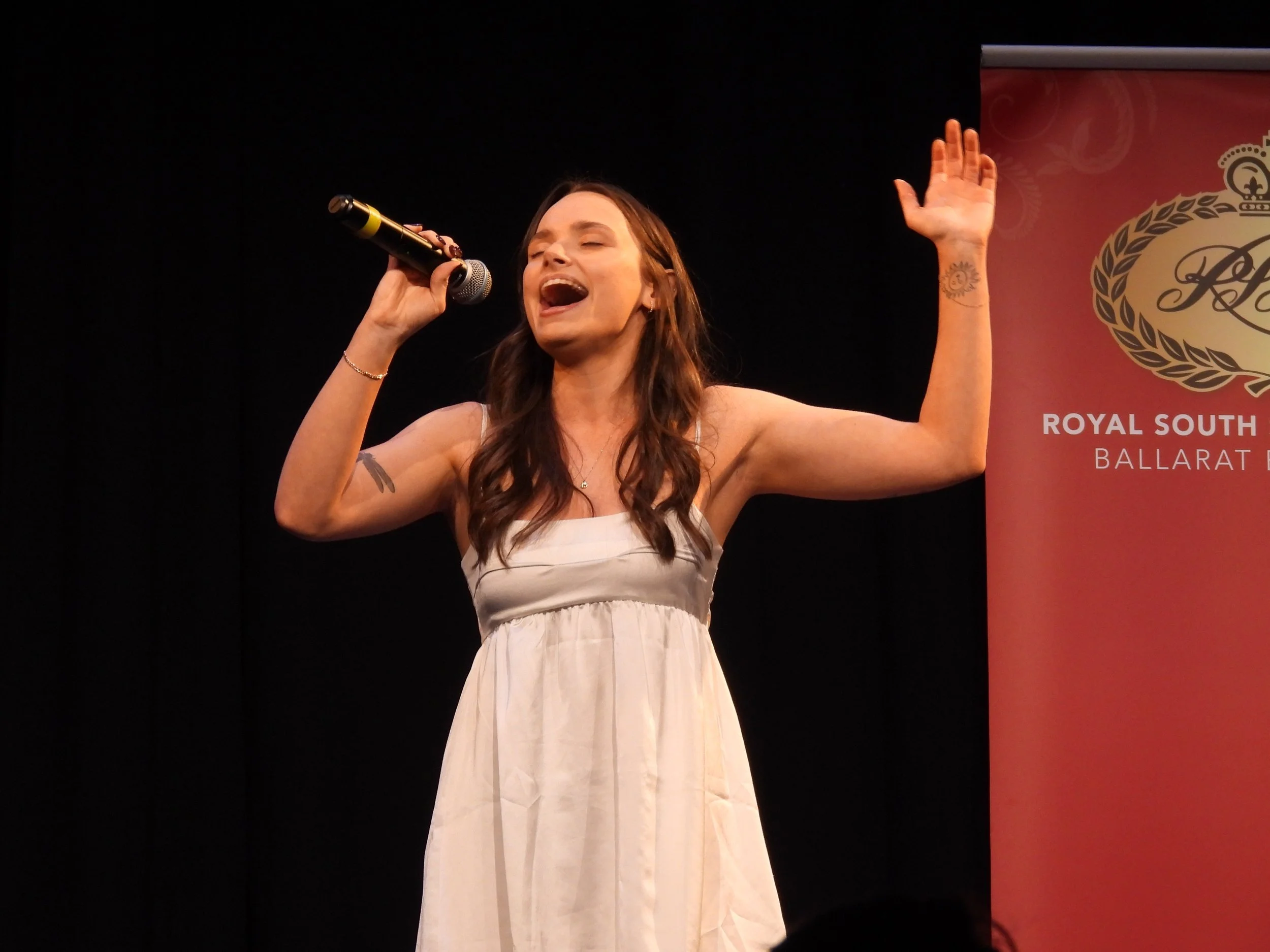 A woman singing into a microphone on stage, with one arm raised and eyes closed, next to a banner with a crest and text that reads "Royal South Street Ballarat."