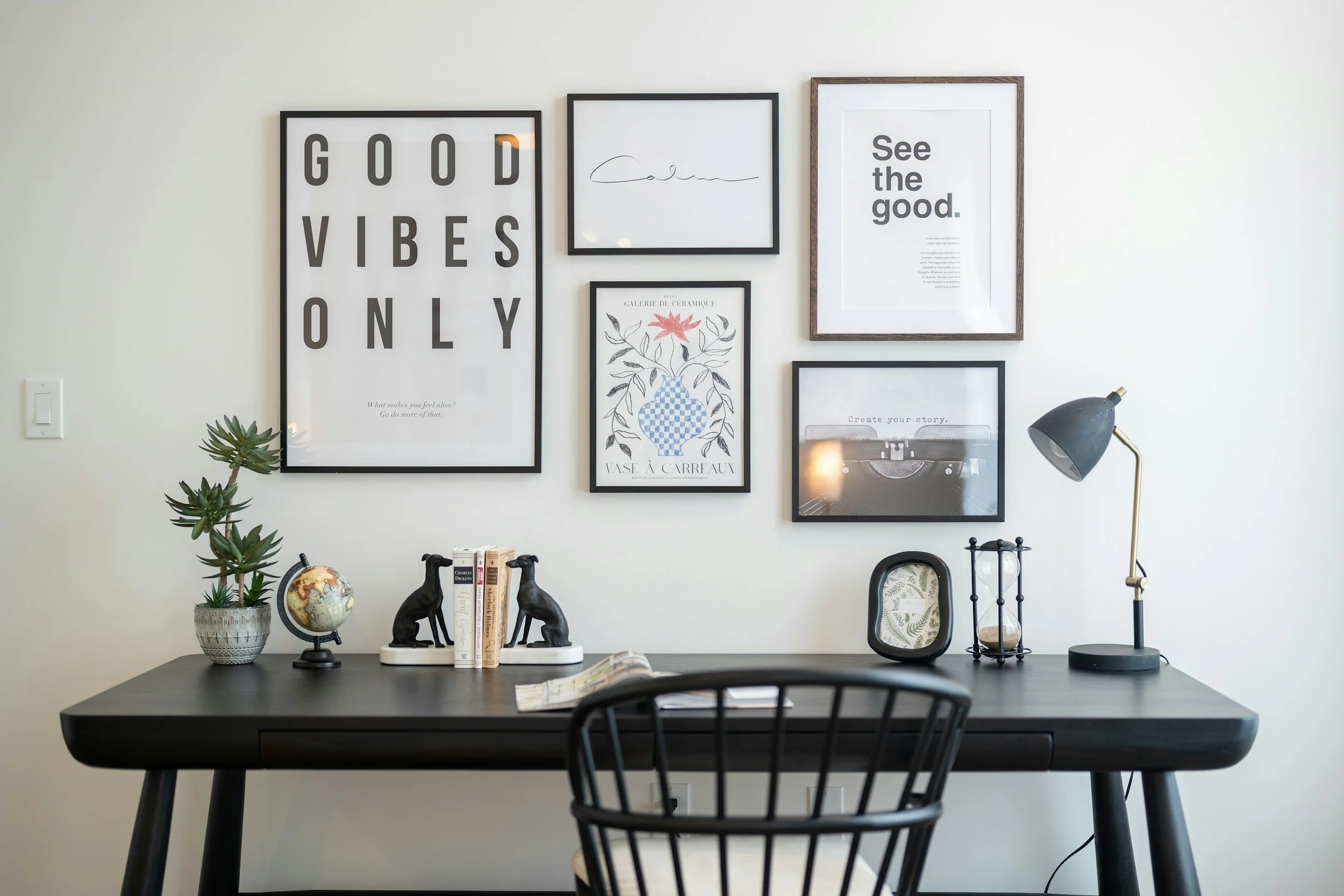 A modern home office workspace with framed wall art on a white wall, a black desk, a chair, a black desk lamp, a small globe, and decorative items including a succulent plant and dog figurines.