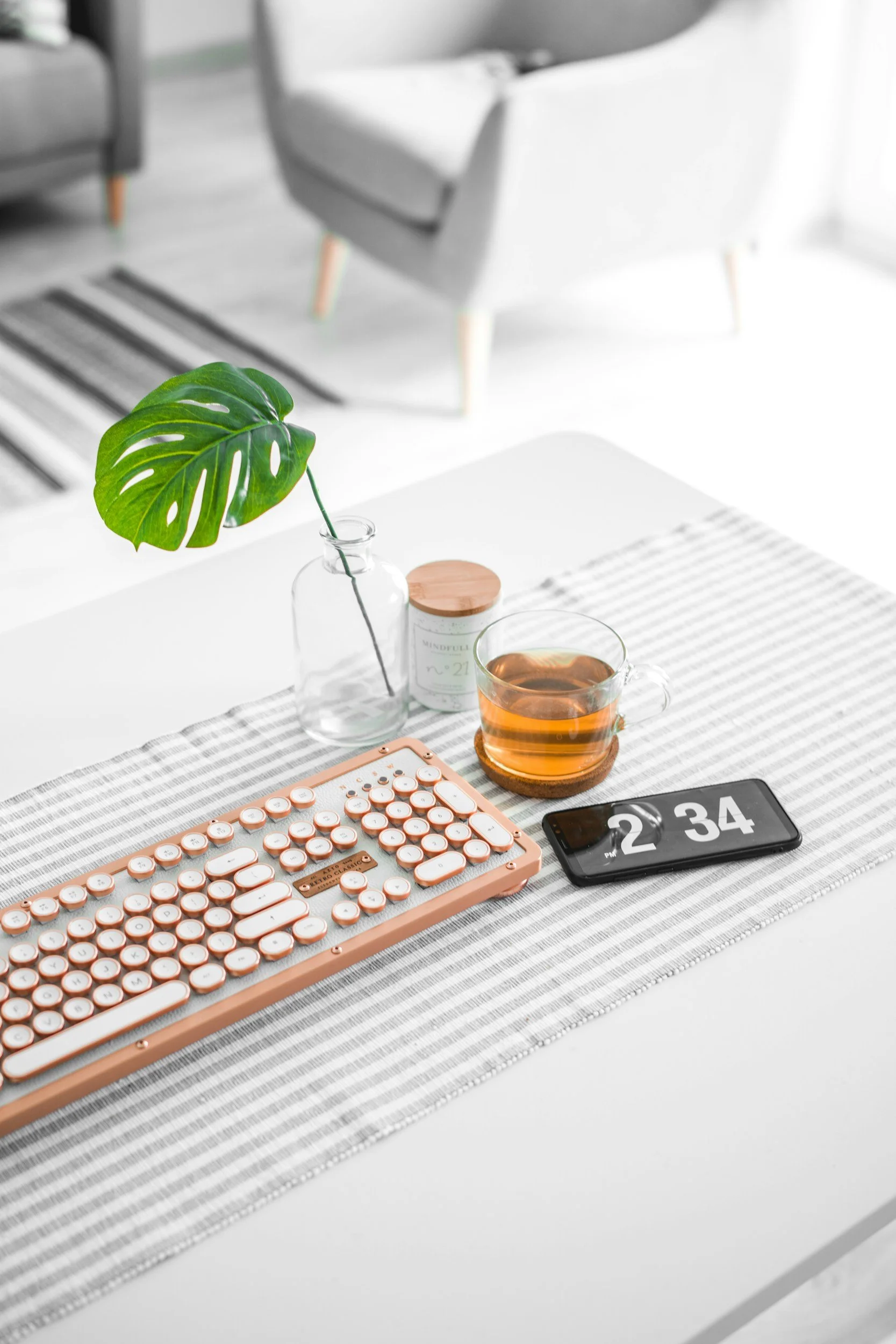 A white table with a striped table runner holds a pink keyboard, a bottle with a green Monstera leaf, a candle, a glass cup of tea, a smartphone displaying 2:34, and a small pink device.