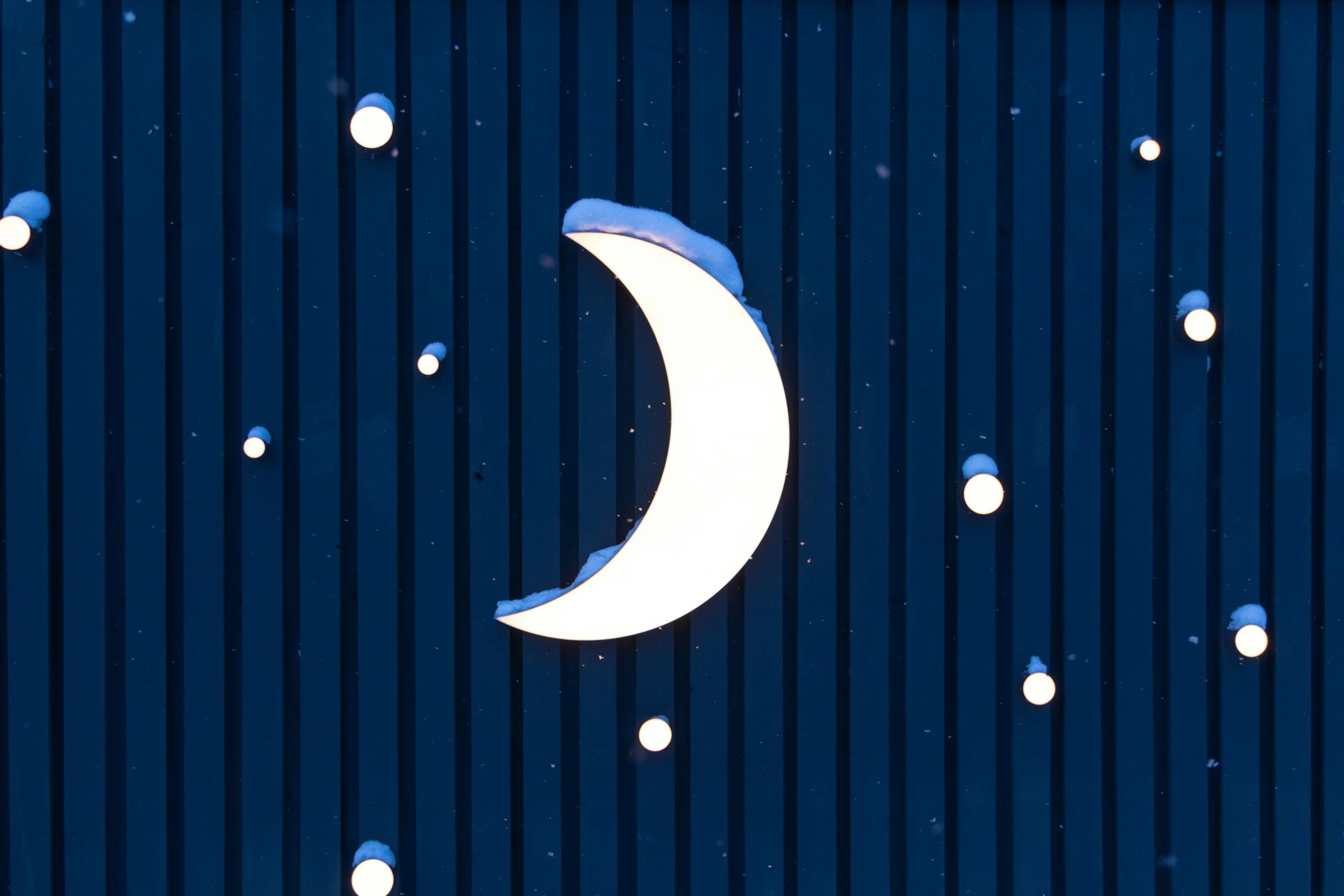 A crescent moon-shaped light fixture with snow on top, mounted on a dark striped wall, with small round lights surrounding it.