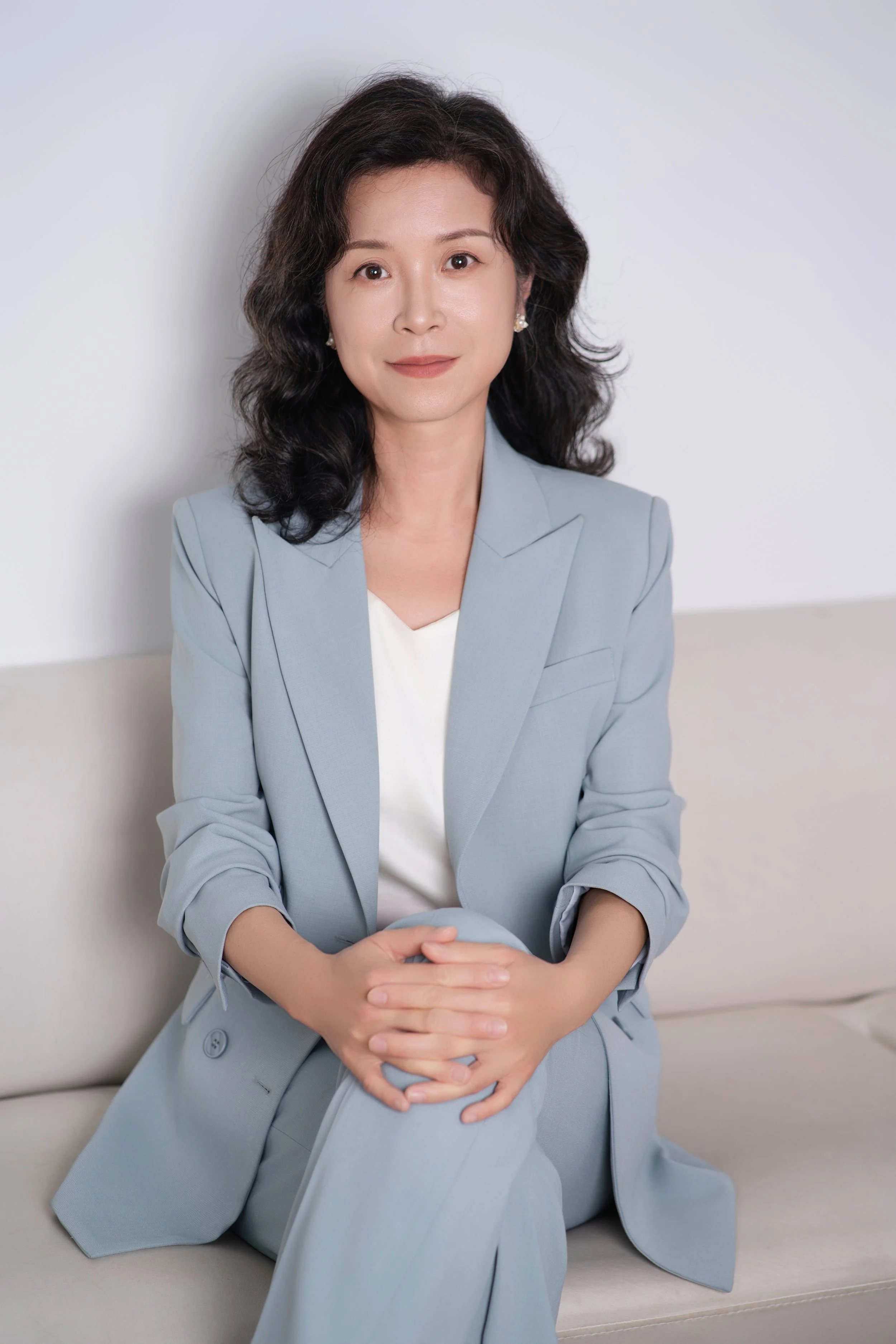 A woman with wavy dark hair and earrings sitting on a beige sofa, wearing a light blue blazer and matching pants, with her hands clasped together in her lap, looking at the camera against a plain light background.