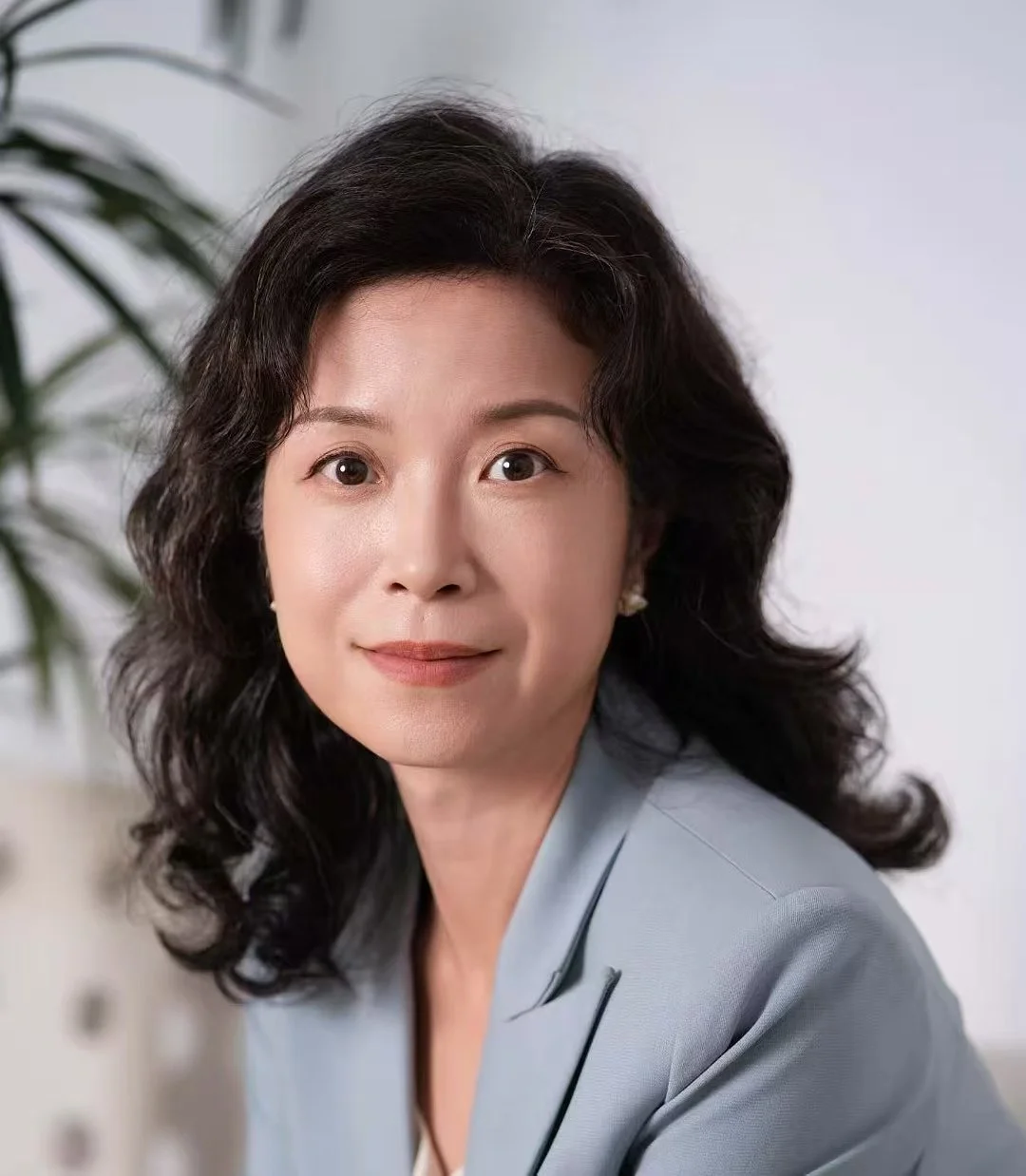 Portrait of a middle-aged woman with black, wavy hair, wearing a light gray blazer, smiling gently at the camera, with a white wall and a plant in the background.