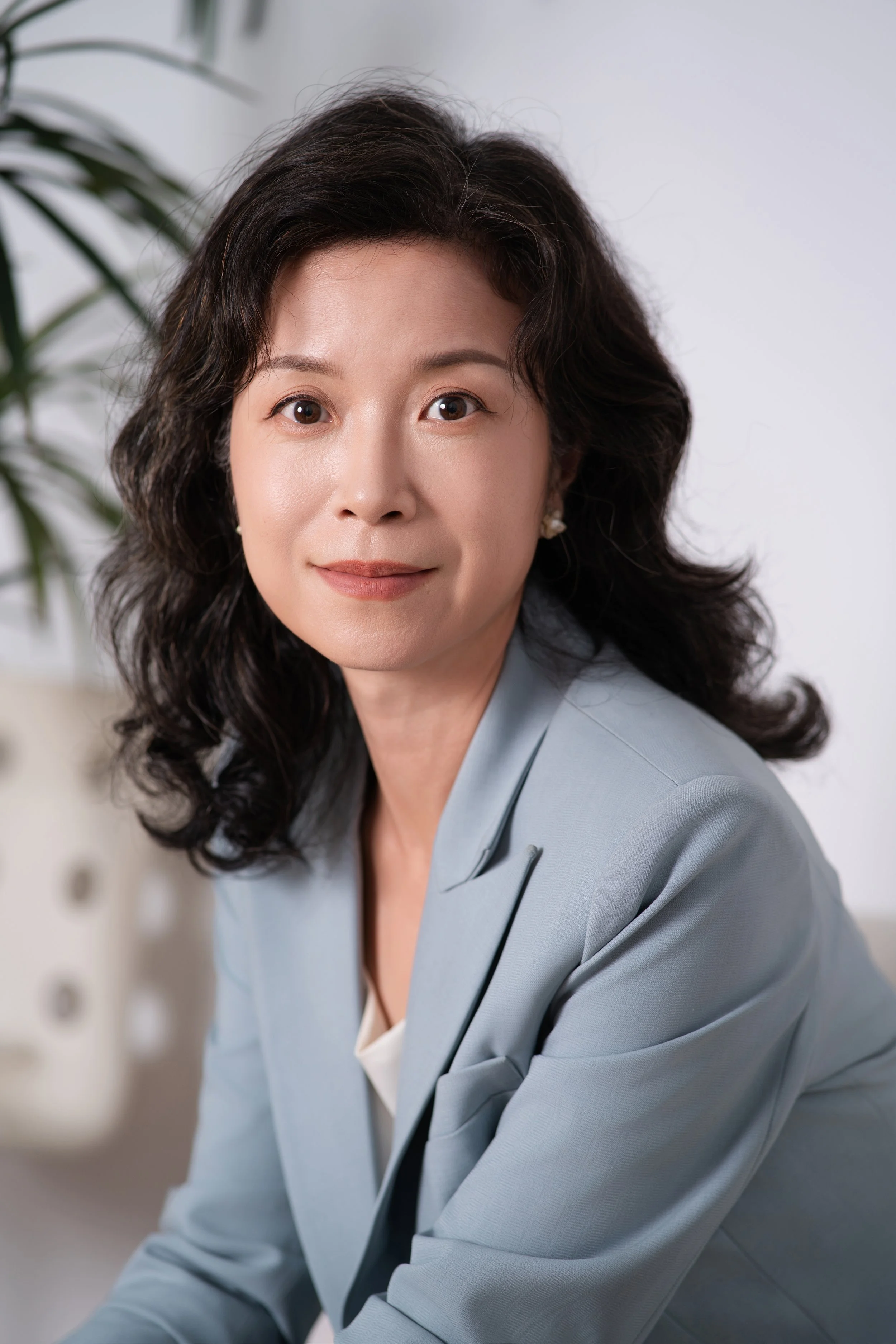 Close-up photo of a middle-aged woman with dark, wavy hair, wearing a gray blazer and earrings, smiling subtly, in an indoor setting with a plant in the background.