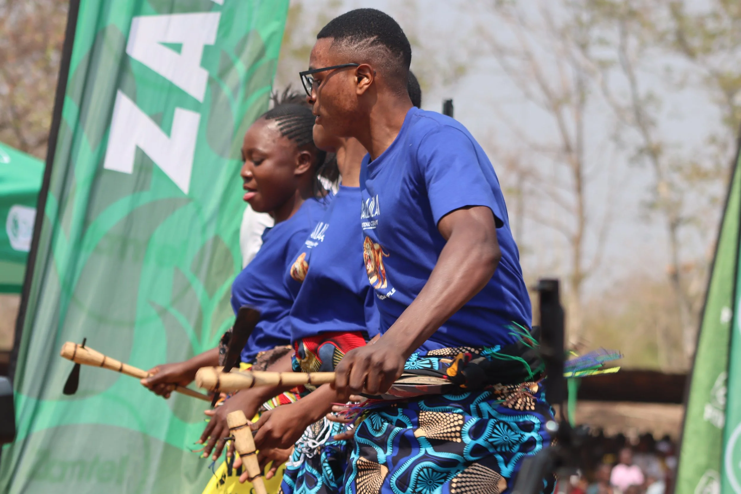 Two individuals are participating in a traditional dance or performance outdoors, holding wooden sticks, with trees and green banners in the background.