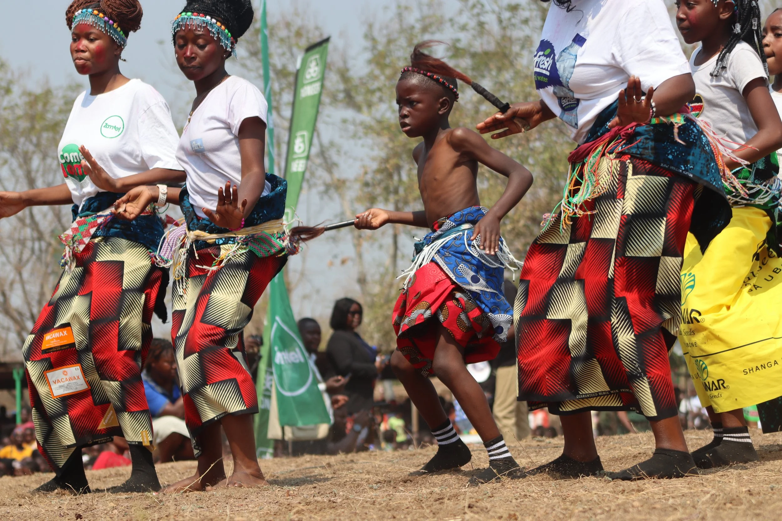 A group of people, including children, are participating in a traditional dance outdoors. They are dressed in colorful clothing, some are wearing beaded accessories and headbands. The children are barefoot or wearing simple footwear, and there are gr