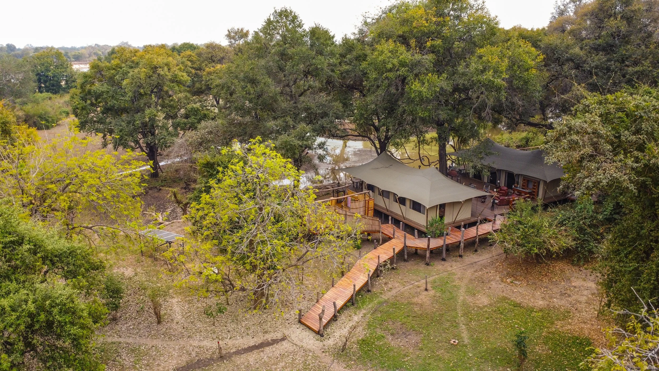 Aerial view of a riverbank with trees and a wooden deck, featuring a large tent and outdoor furniture, in a natural setting.