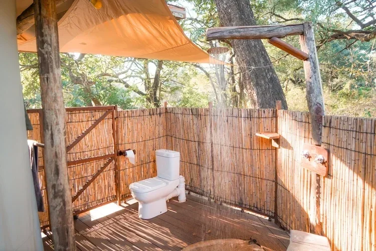 An outdoor bathroom with a toilet, enclosed by bamboo fencing, with trees in the background and a canopy overhead.