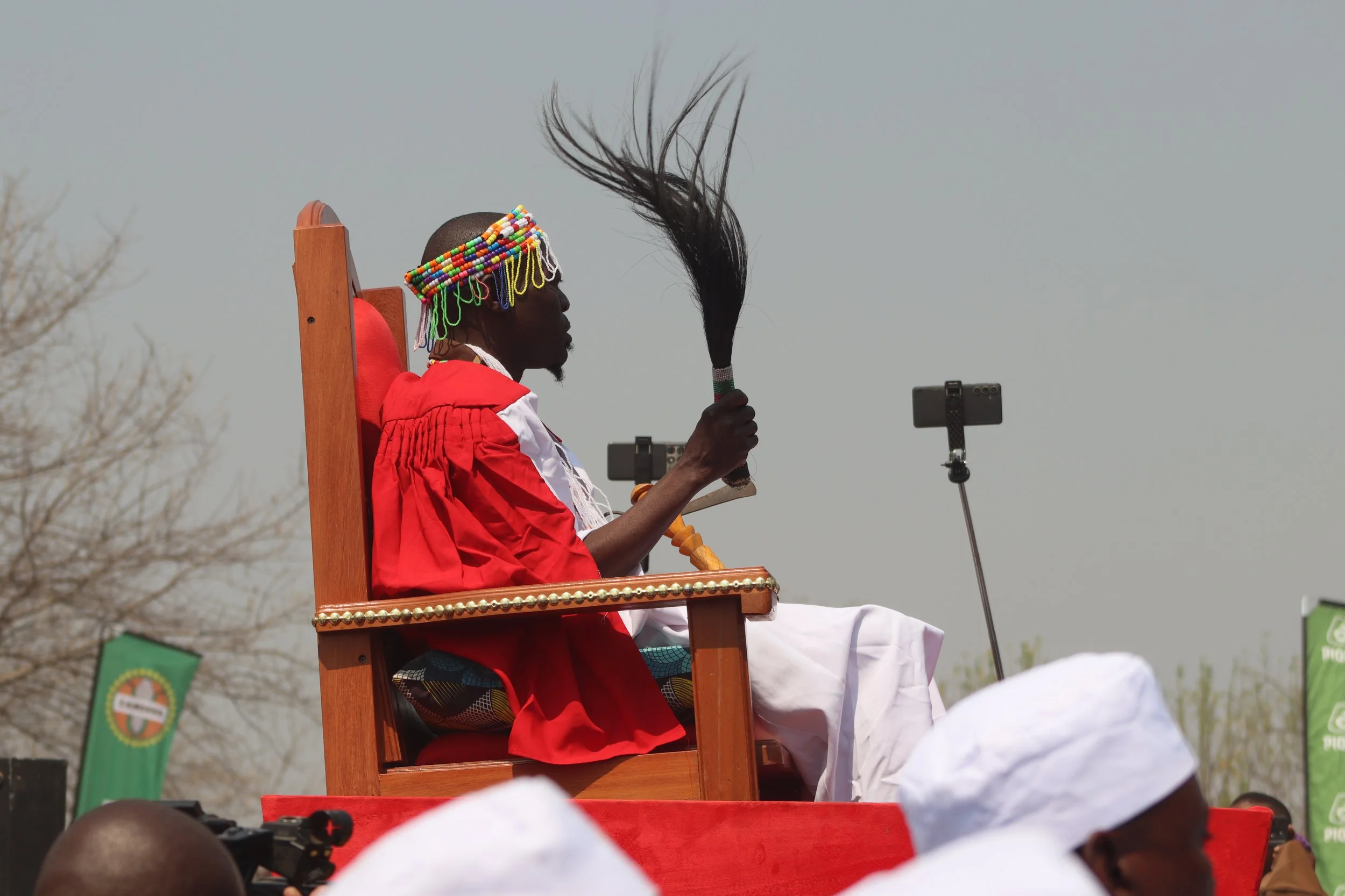 A person wearing traditional Maasai attire and a beaded headdress, sitting on a wooden chair, holding a large brush, with a microphone stand nearby during an outdoor event with audience members.