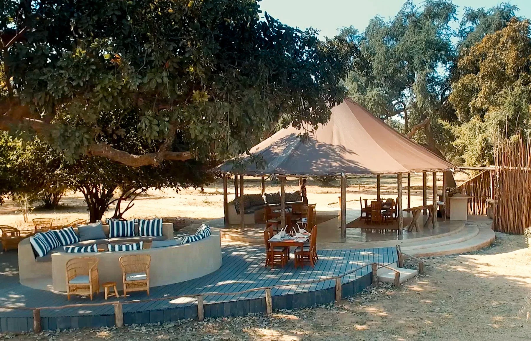 Outdoor seating area with a circular sofa with striped cushions, a dining table with chairs, and a shaded structure under trees, on a wooden deck in a natural setting.