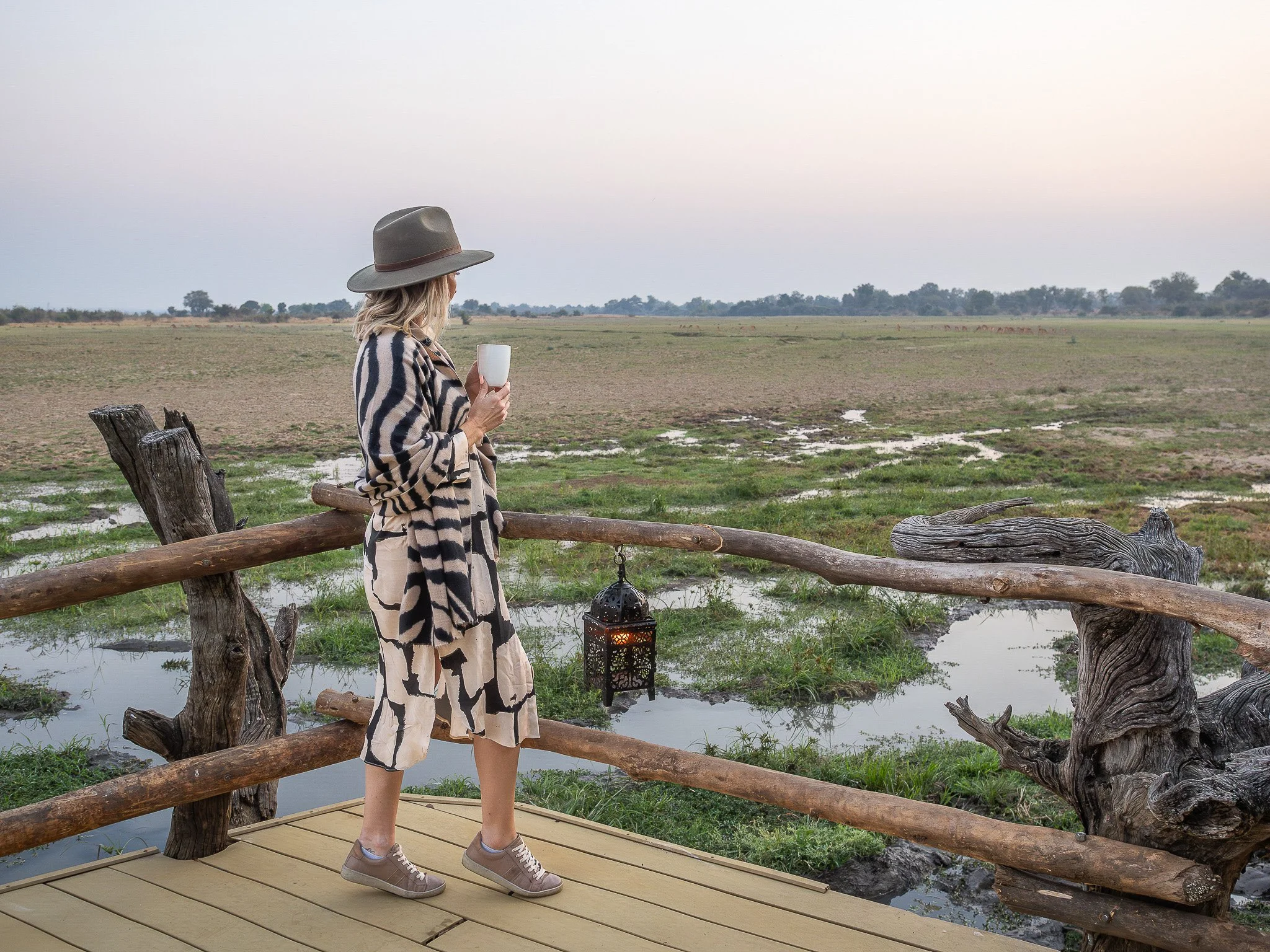 A woman standing on a wooden platform near water, holding a white mug, wearing a large hat, striped coat, and beige pants, looking out over a rural landscape with trees and open fields.