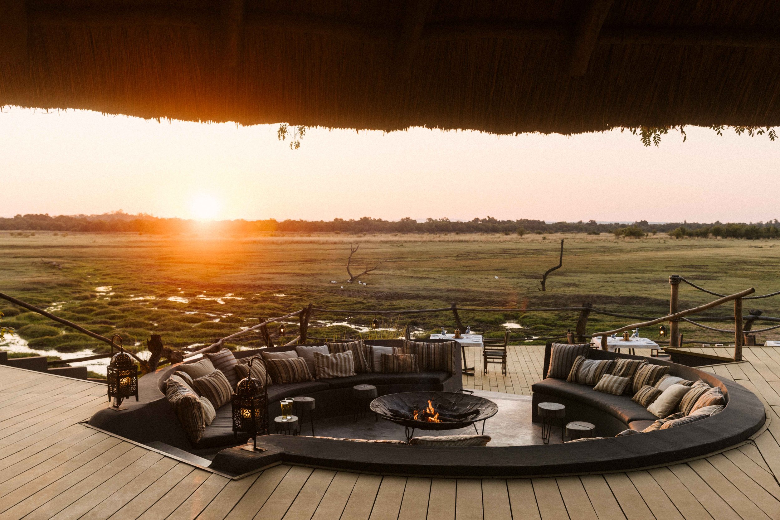 An outdoor seating area with cushioned furniture around a fire pit, overlooking a grassy landscape at sunset.
