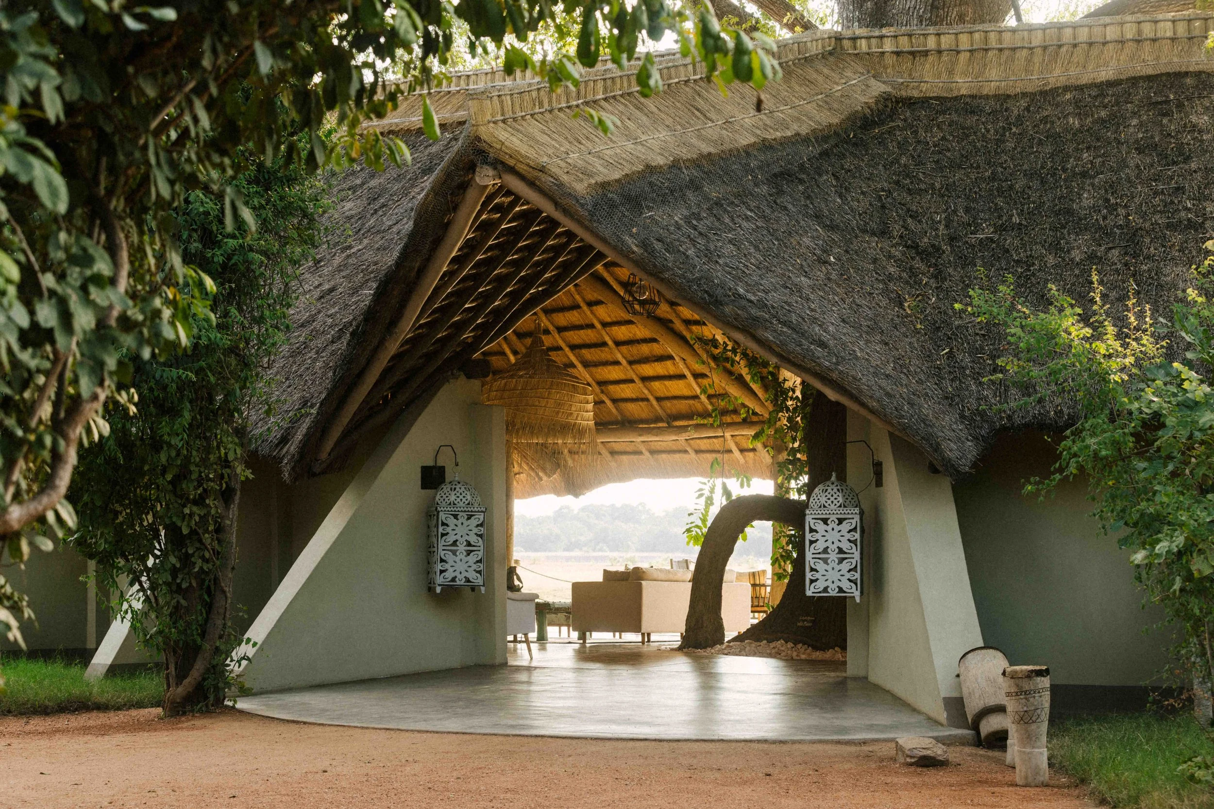 A thatched roof pavilion with decorative lanterns on white walls, surrounded by greenery and trees.