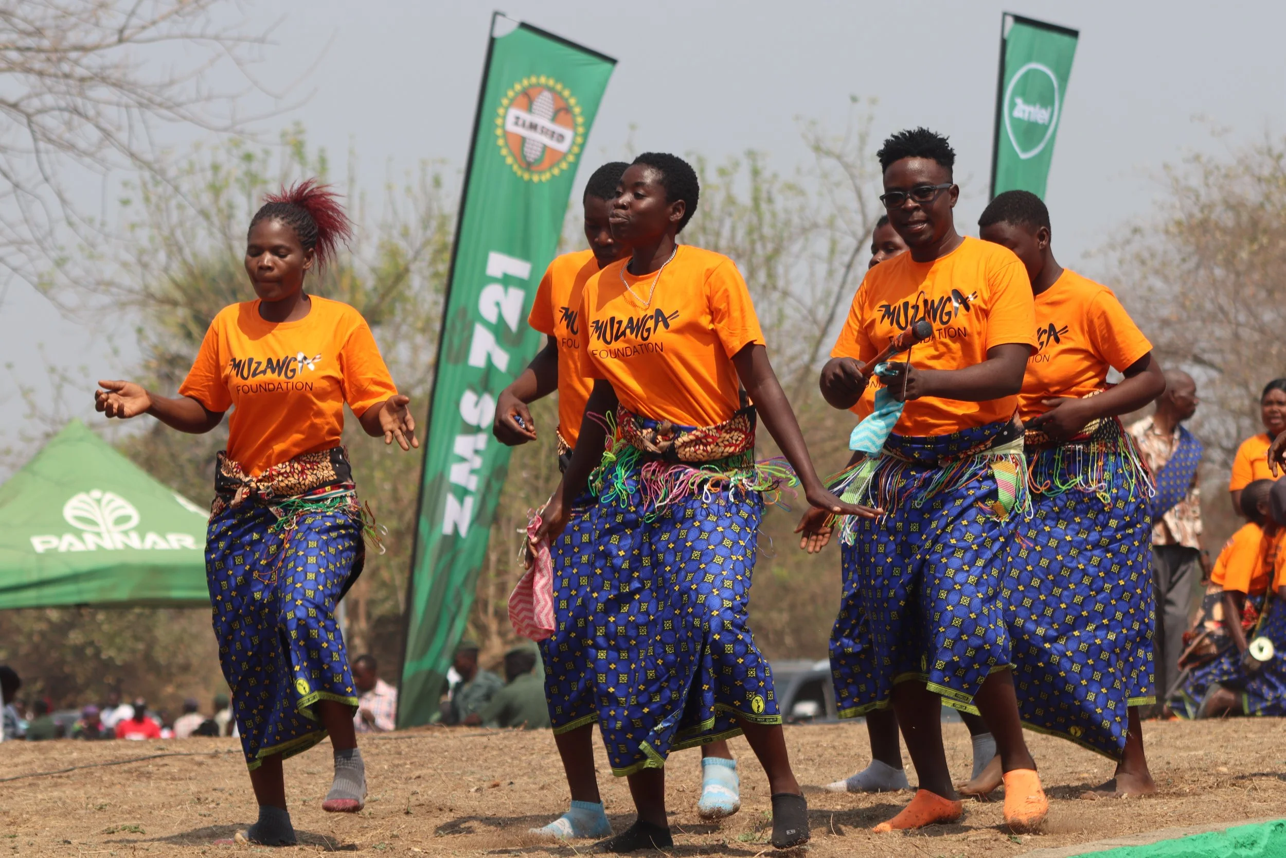 Group of women dancing outdoors at an event, wearing matching orange T-shirts with the words 'Muzanga Foundation,' and colorful patterned skirts. There are banners in the background with logos and text, and other people can be seen in the distance.