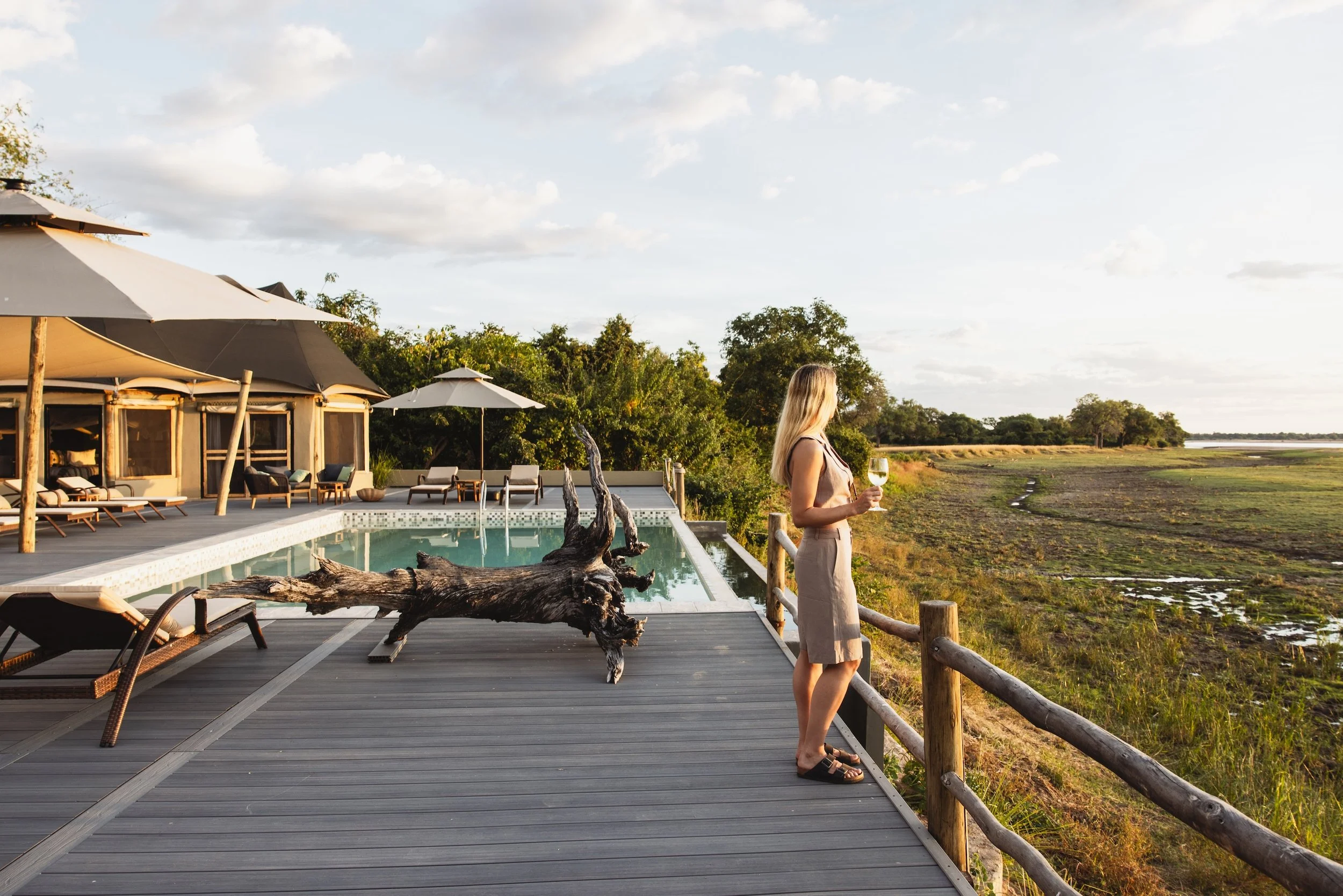 A woman standing on a wooden deck overlooking a marsh with wetlands and trees, holding a glass of white wine, with a hotel or resort building with umbrellas and lounge chairs nearby, during the late afternoon or early evening.