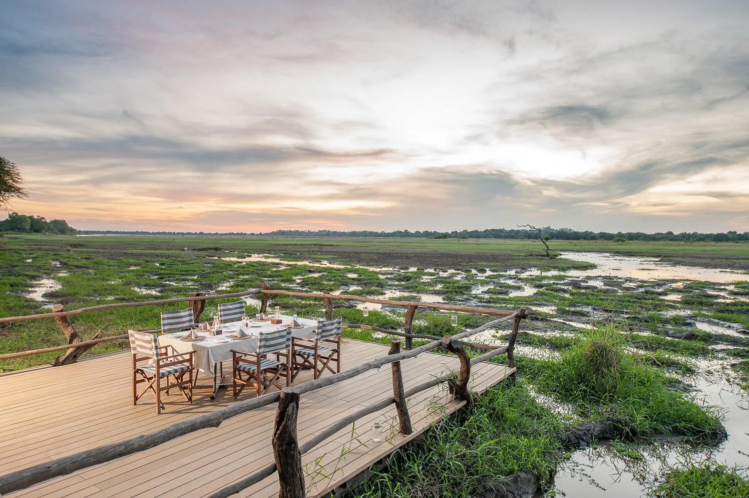 Dining table set on a wooden deck overlooking a wetland at sunset with cloudy sky.