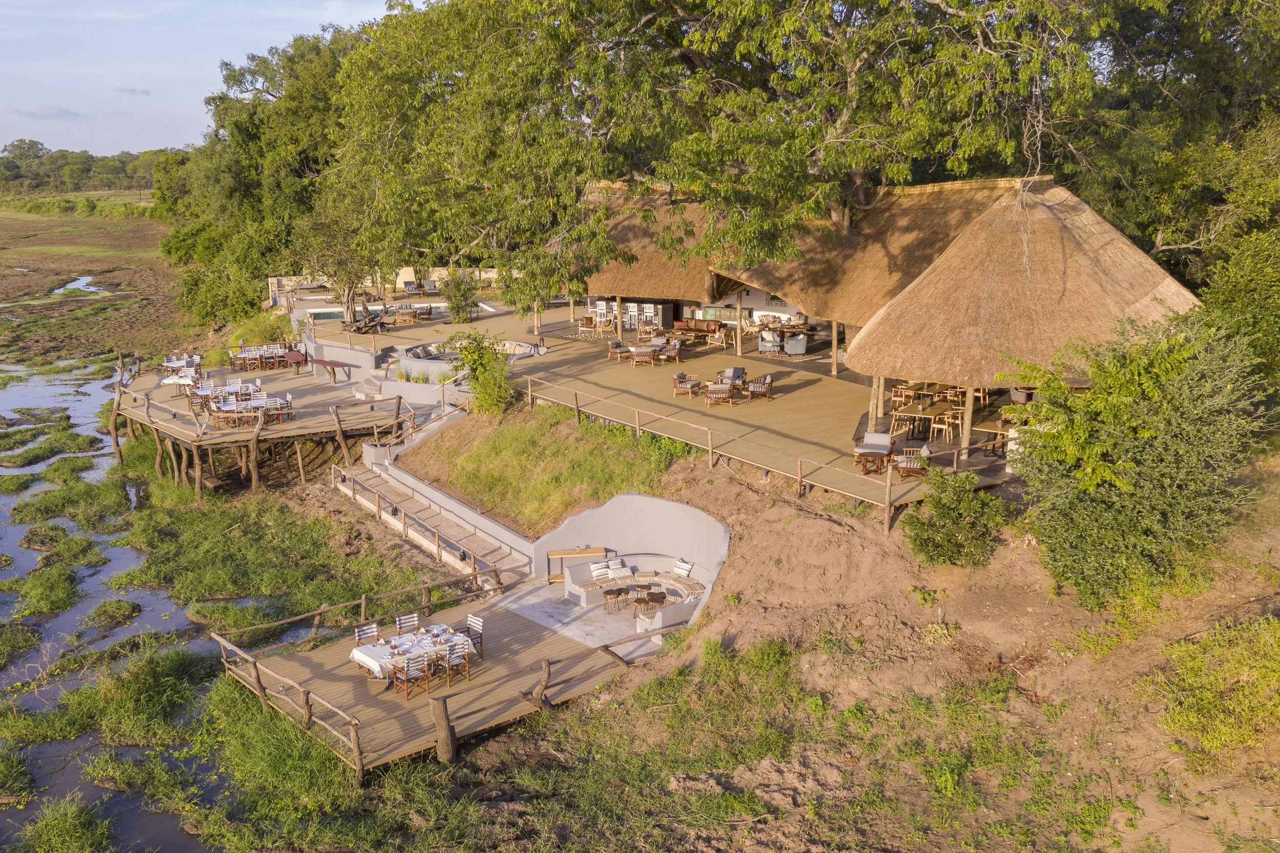 Aerial view of an outdoor dining and lounge area with thatched-roof structures, surrounded by lush greenery and water features, in a natural setting.