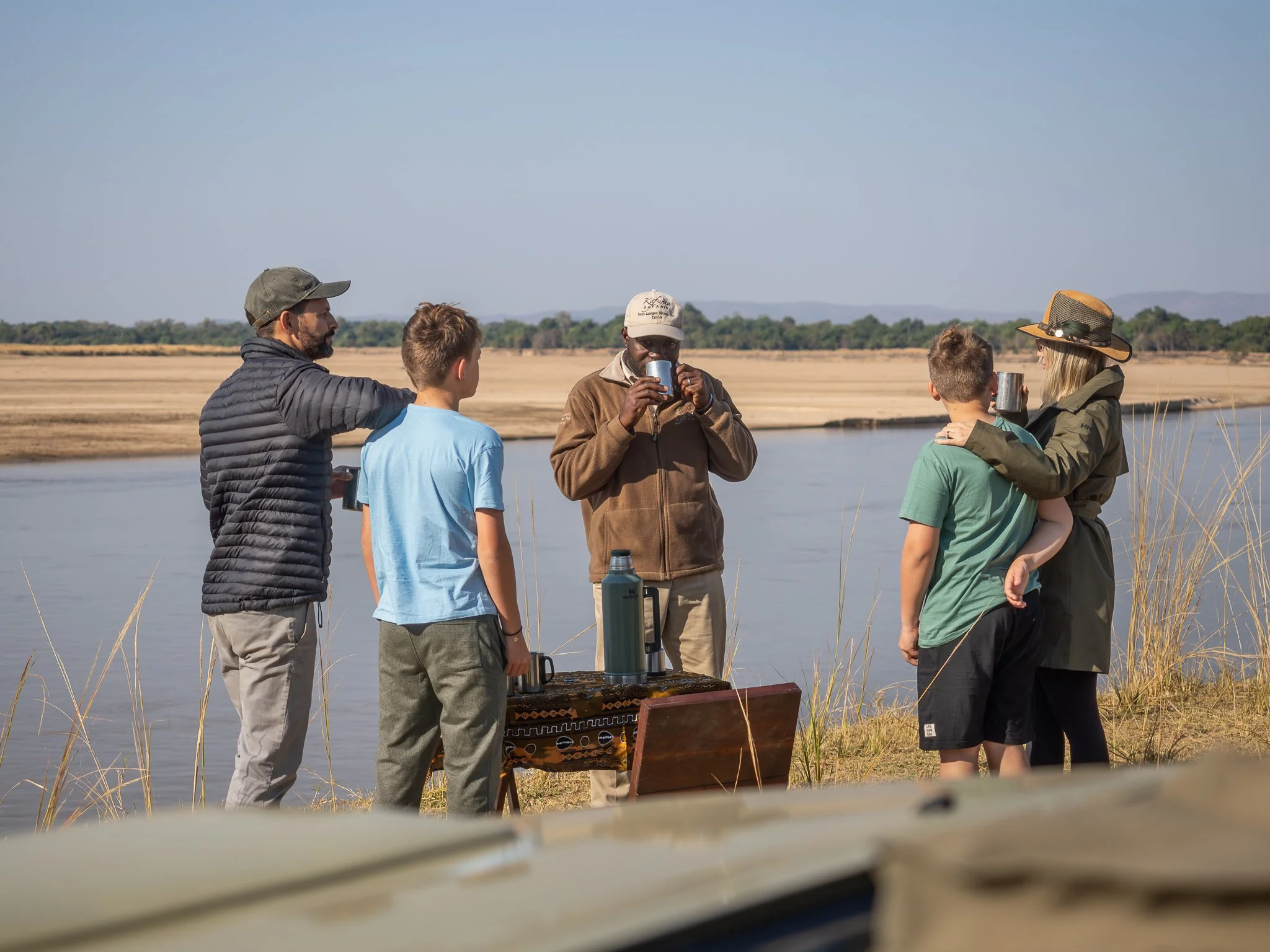 Group of people standing outdoors by a body of water, enjoying drinks and conversation on a sunny day.