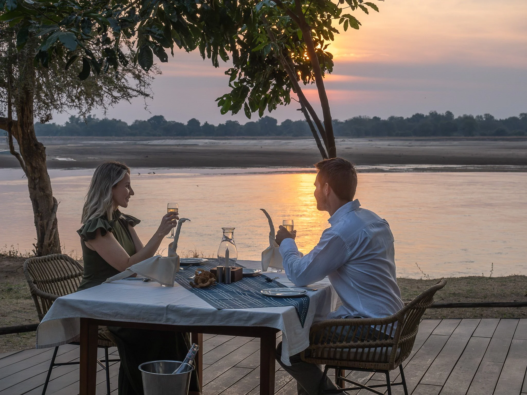 A couple enjoying a meal outdoors at a restaurant by a body of water during sunset. The scene includes a table with a tablecloth, wine glasses, and a basket of bread, with trees and the setting sun in the background.