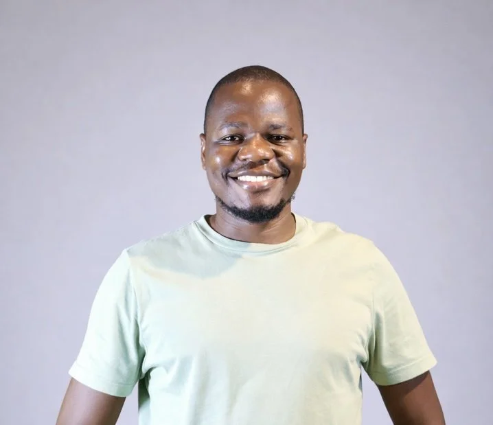 A smiling man with a short beard, wearing a light green t-shirt, standing against a plain gray background.