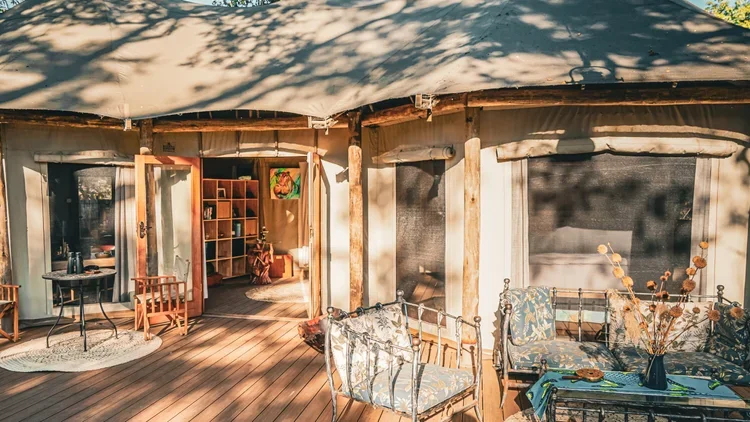 Outdoor patio with wooden flooring, metal chairs with floral cushions, a table with a blue vase and dried flowers, and an open indoor space with shelves, chairs, and a rug.
