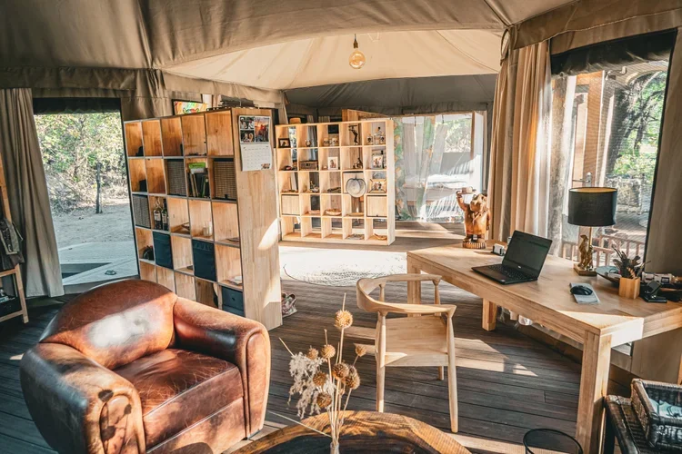 Interior of a cozy tent with wooden furniture, a leather armchair, a desk with a laptop, bookshelf, and decorative items, with sunlight streaming in through the windows.