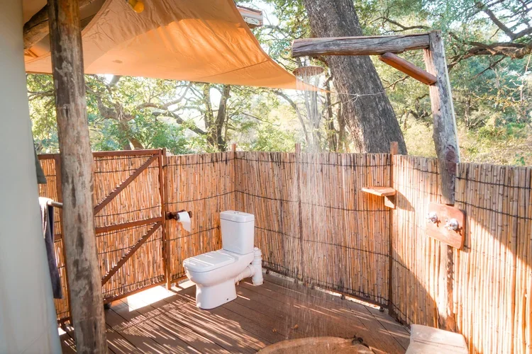 An outdoor bathroom with a white toilet, bamboo privacy fence, shower head, and a small shelf, set in a natural wooded area with trees and sunlight filtering through.