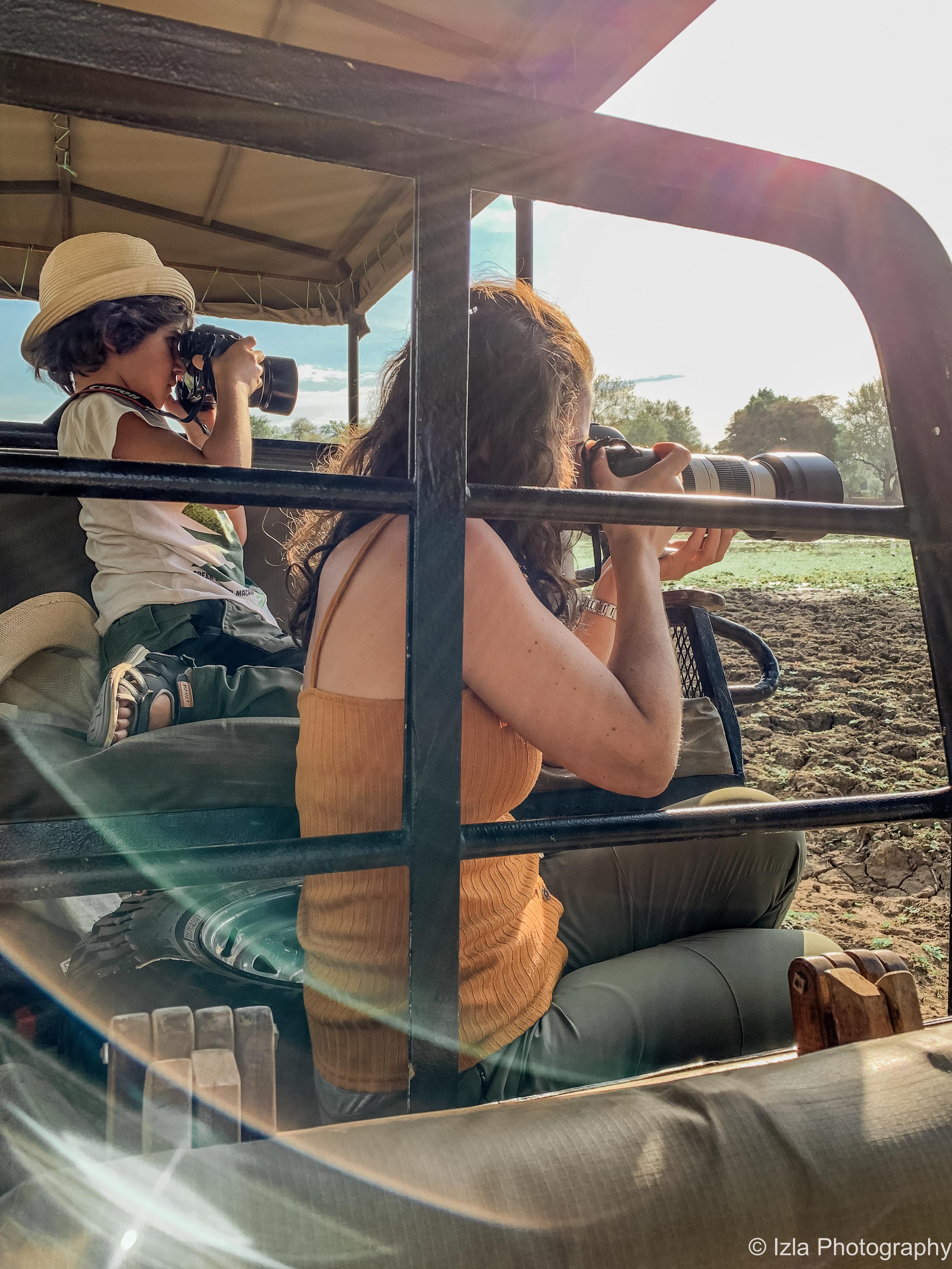 A woman and a young girl, both holding binoculars, sit in an open safari vehicle with a landscape of trees and open land in the background.