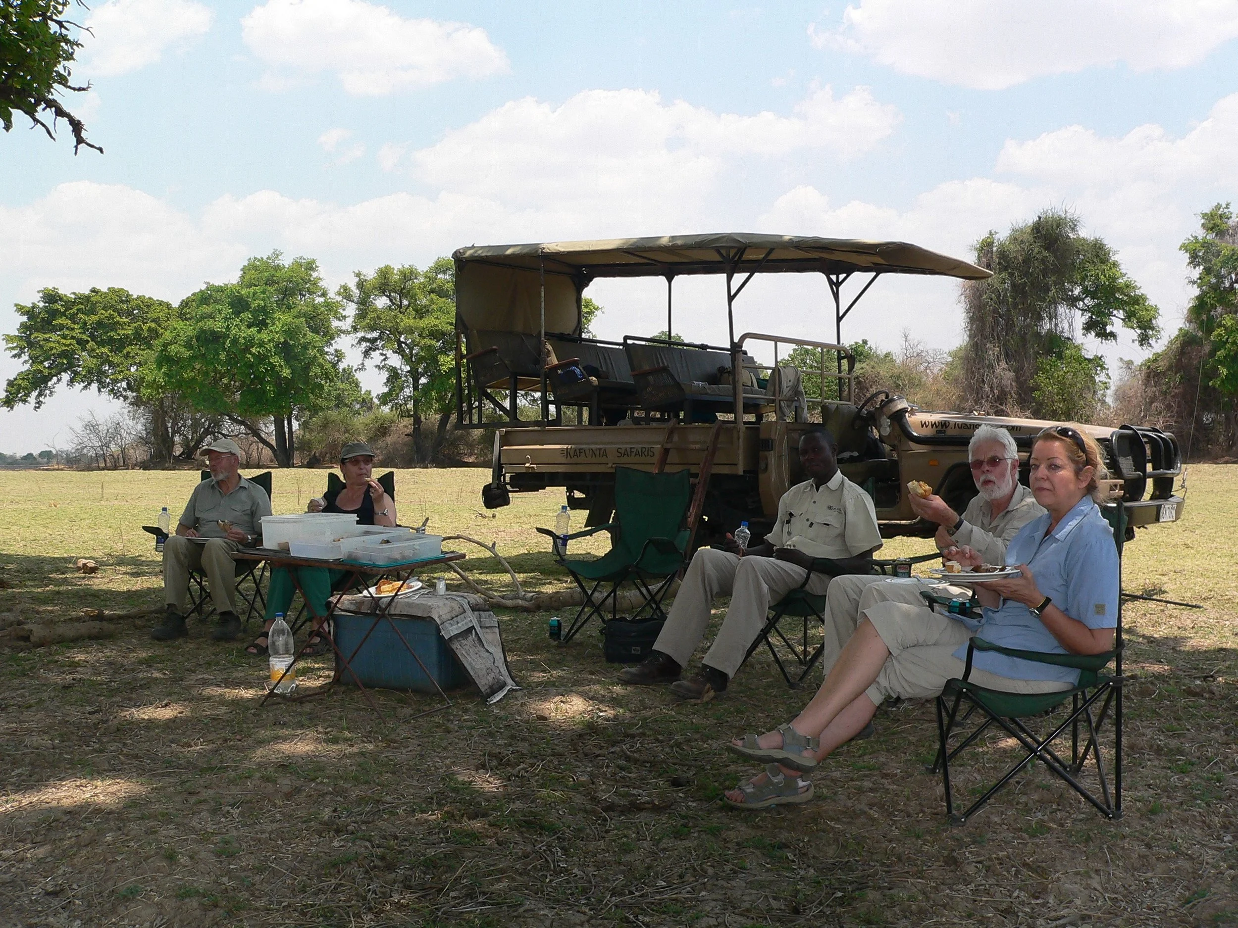 Group of people sitting outdoors on camping chairs under trees, with equipment and food containers nearby, and a safari vehicle in the background during daytime.