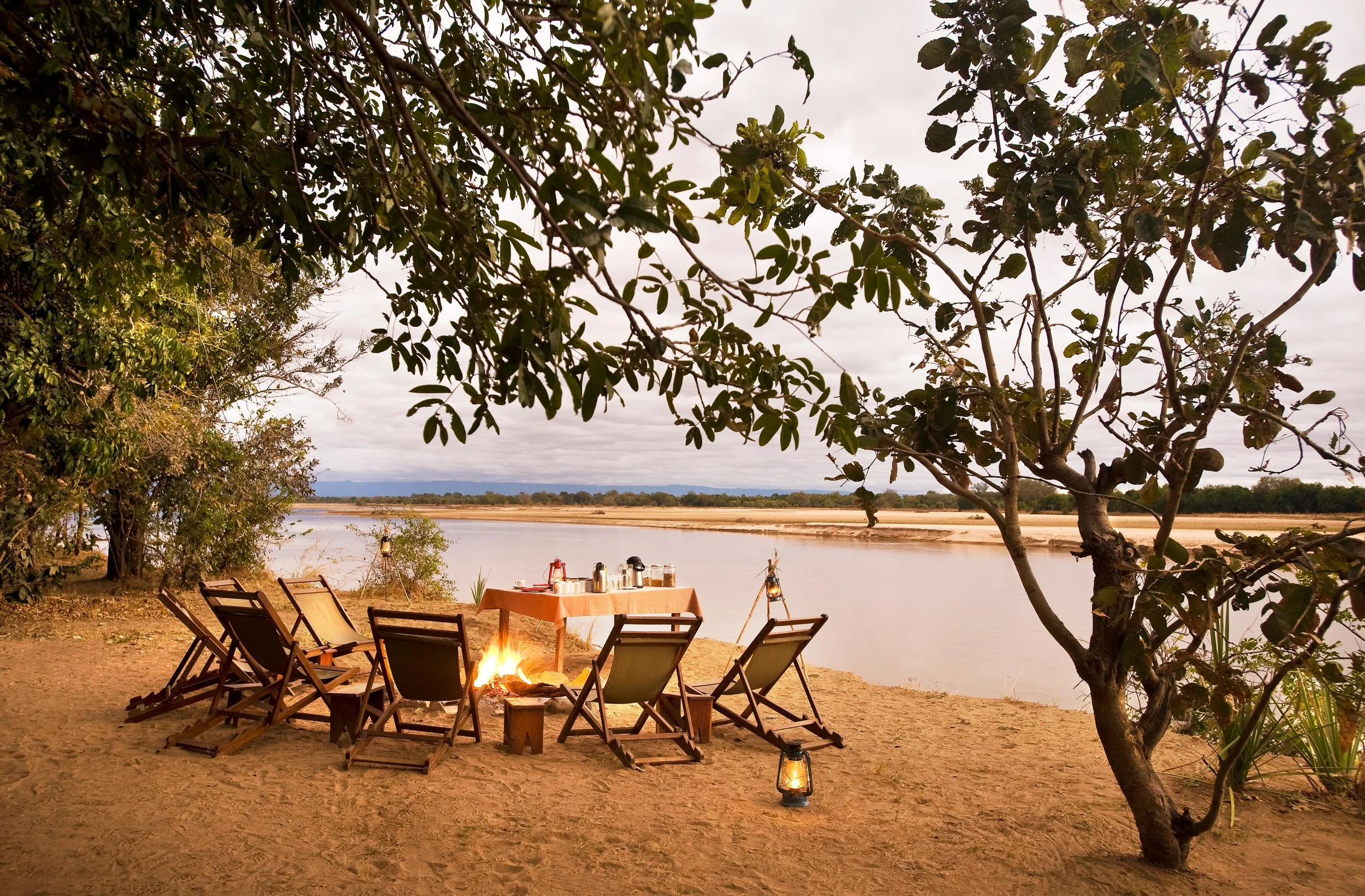 A lakeside outdoor dining setup with a small table covered with an orange tablecloth, surrounded by wooden chairs, a campfire, and lanterns, under a large tree with lush green leaves, overlooking a calm body of water with a wide sandy shoreline and a cloudy sky in the background.