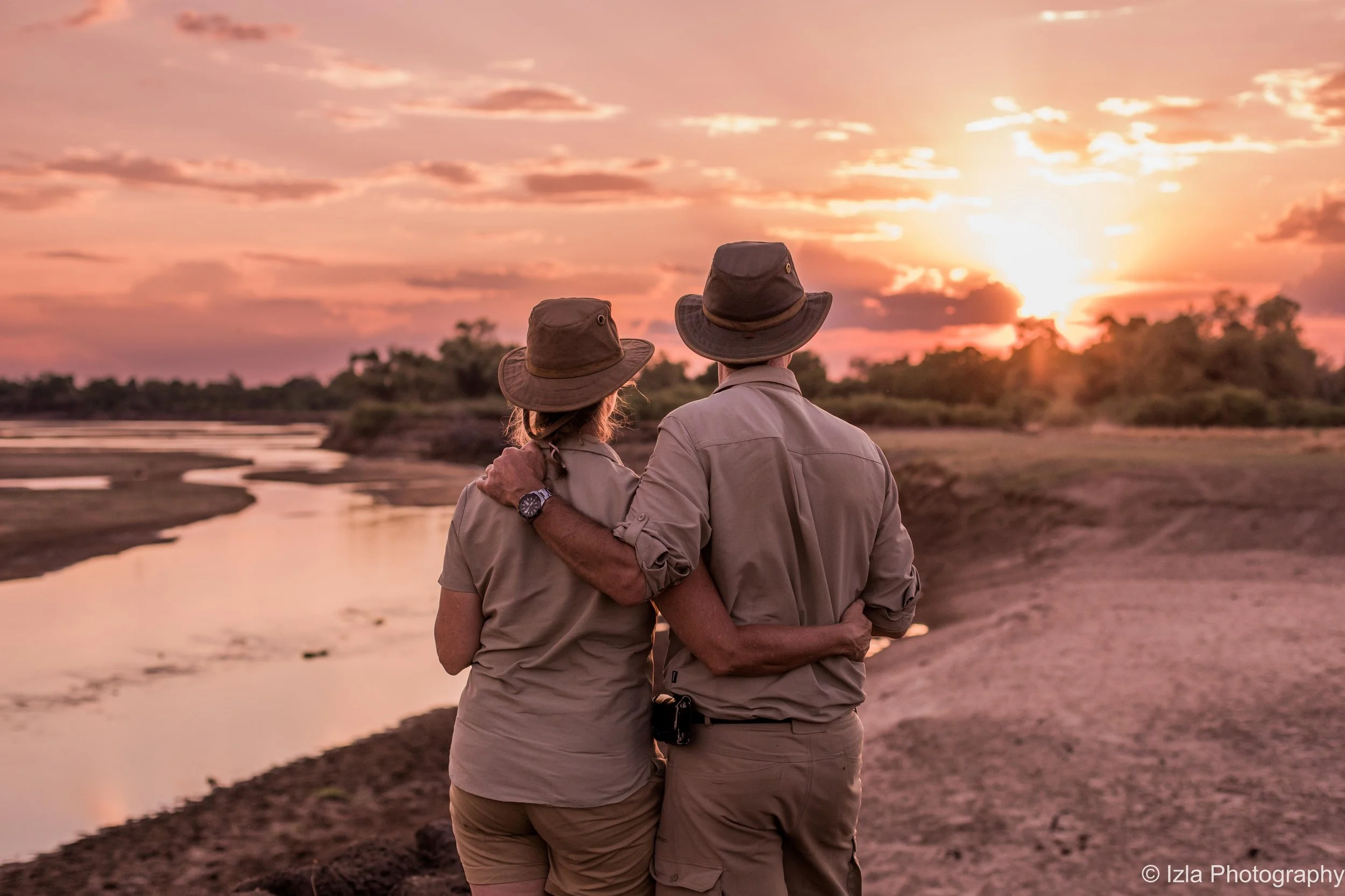 A couple in outdoor explorer clothing, watching the sunset by a river, with their arms around each other.