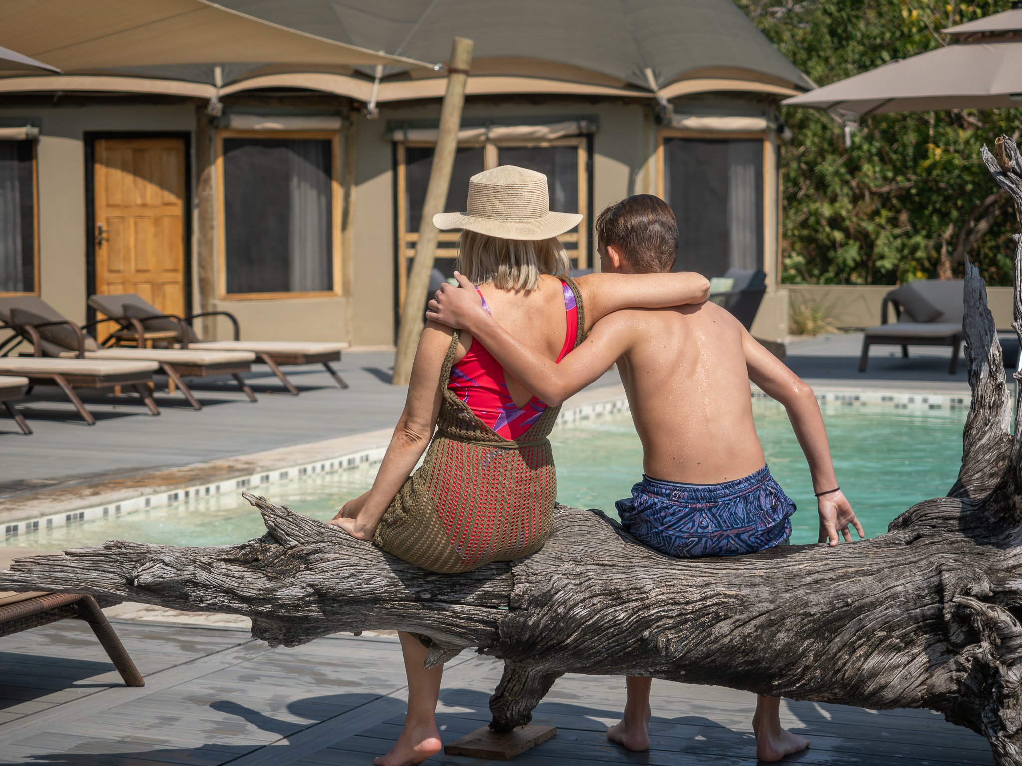 A woman and a man sitting together on a large piece of driftwood at a poolside, embracing each other while facing away from the camera. The woman is wearing a wide-brimmed hat and a colorful swimsuit, and the man is shirtless and wearing blue-pattern
