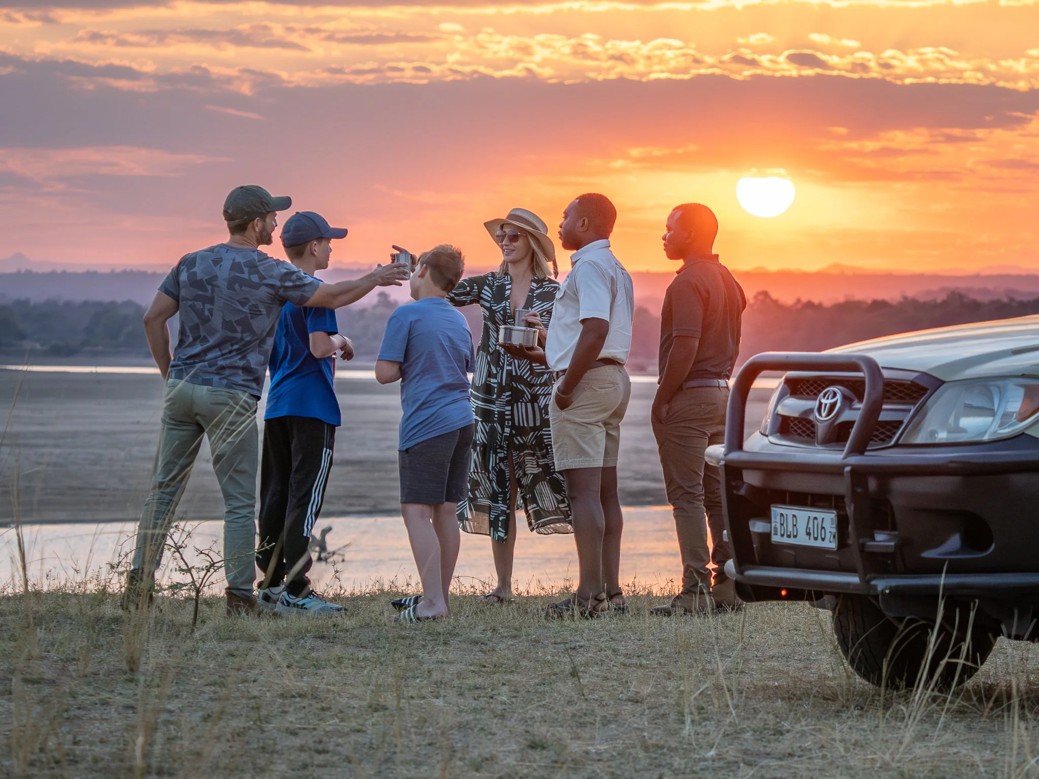Group of six people, including children and adults, standing outdoors near the water at sunset, smiling and taking photos, with cars parked nearby.