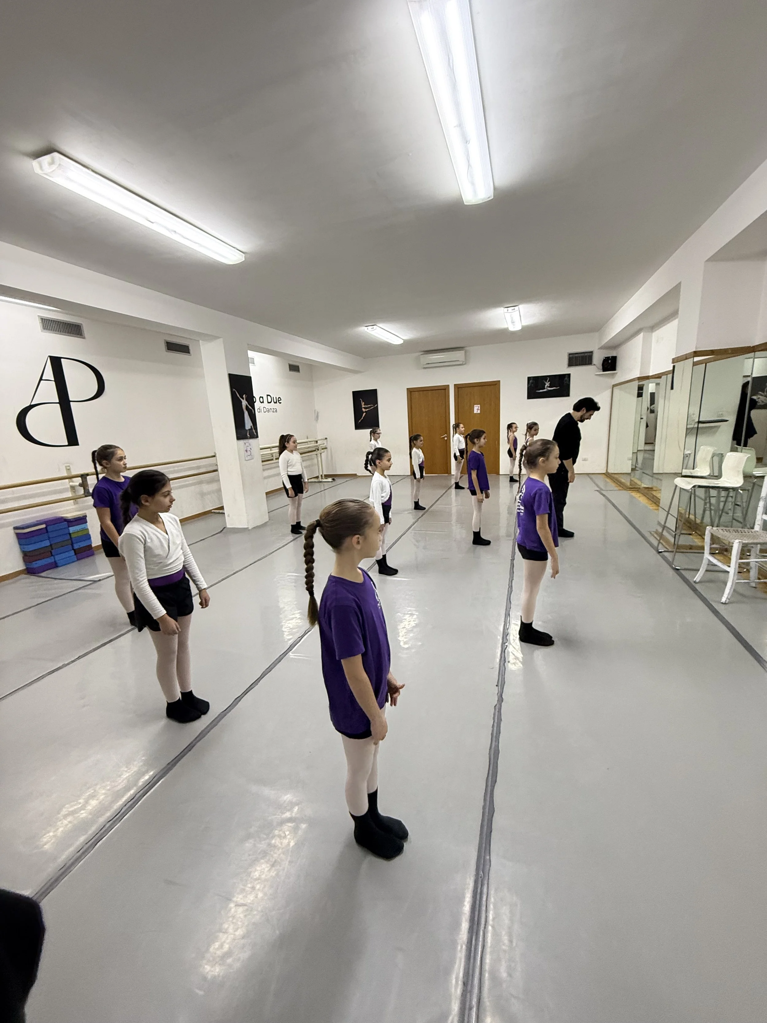 Bambine in una sala di danza, indossano tutù e magliette di diverso colore, si allenano di fronte a uno specchio, con un insegnante che le osserva