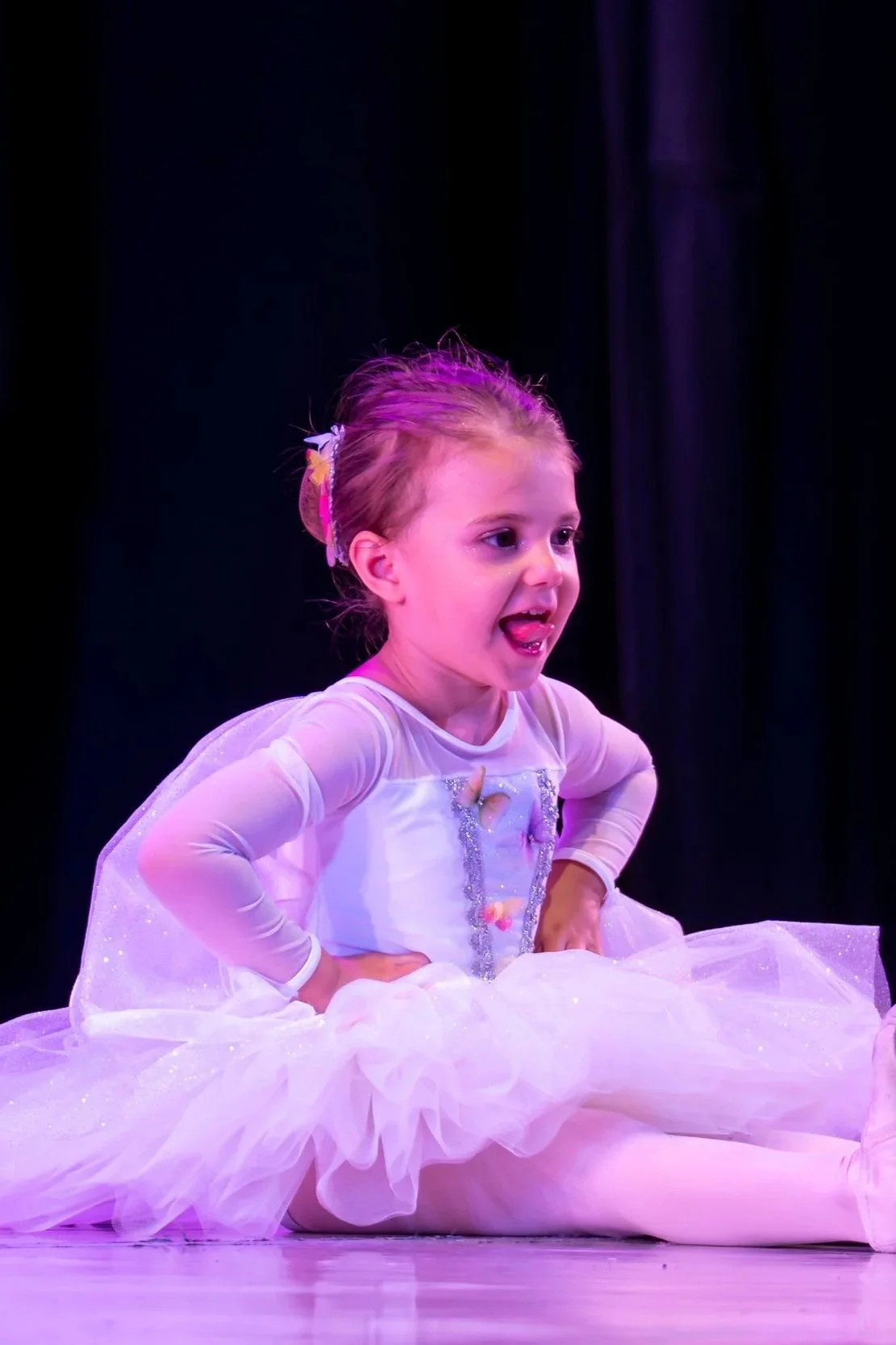 Una giovane ballerina con un costume bianco e gonna di tulle rosa seduta sul palco, con espressione felice e linguetta fuori, sotto luci rosa e viola.