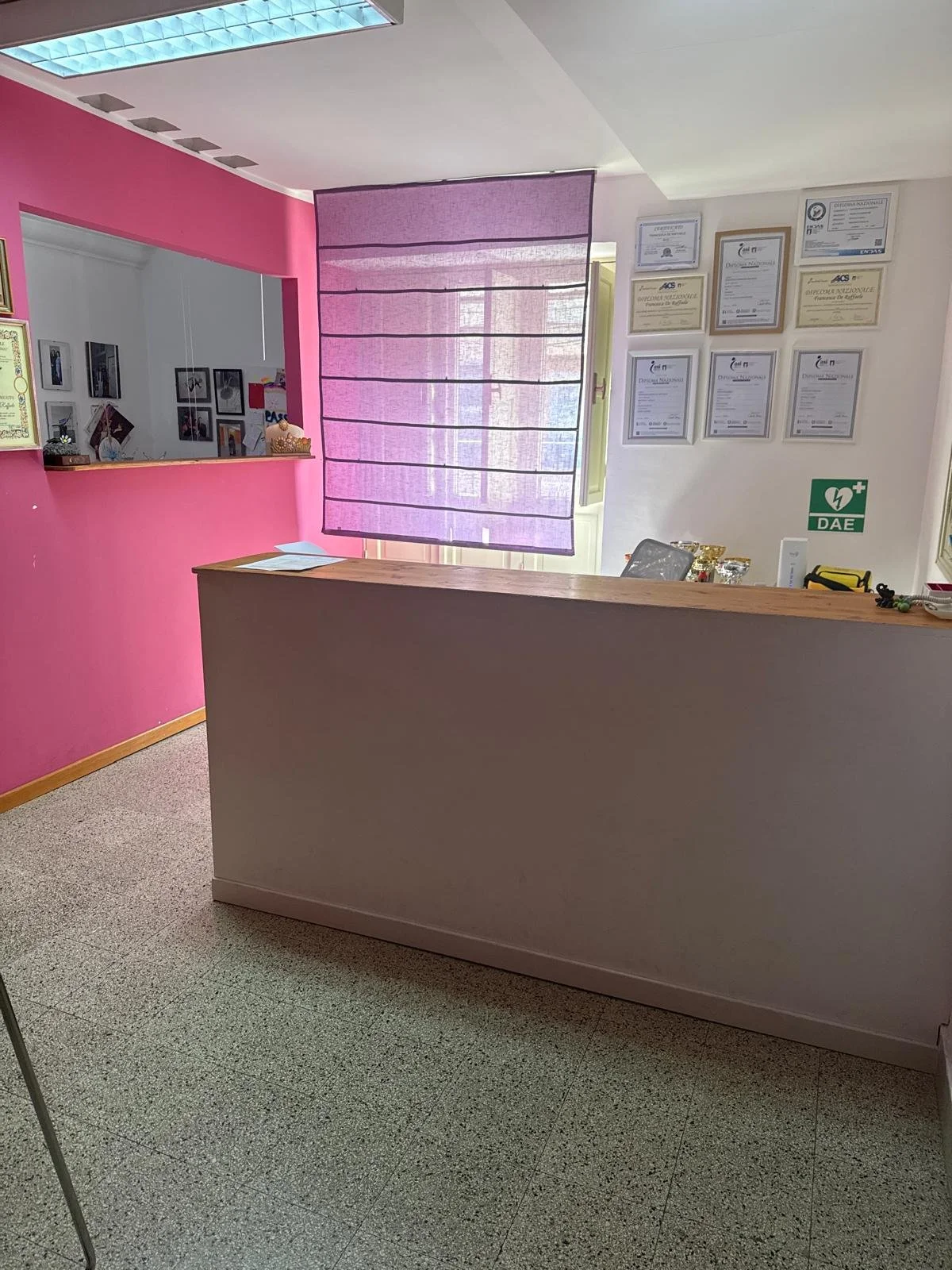 Reception area with a pink wall, purple window blind, framed certificates on white wall, and a wooden counter.