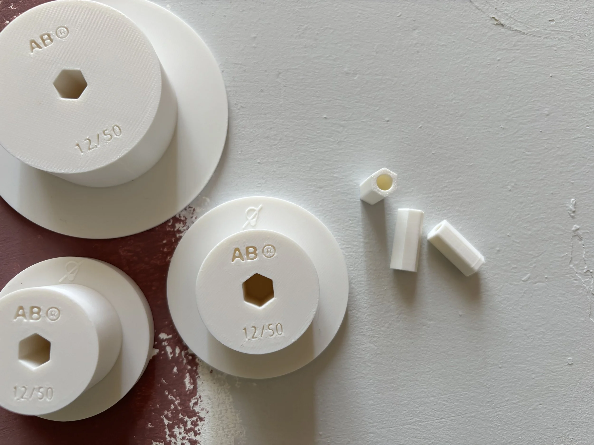 Piastre di plastica bianche con fori centrali e scritte come 'AB' e '12/50' appoggiate su una superficie bianca.
