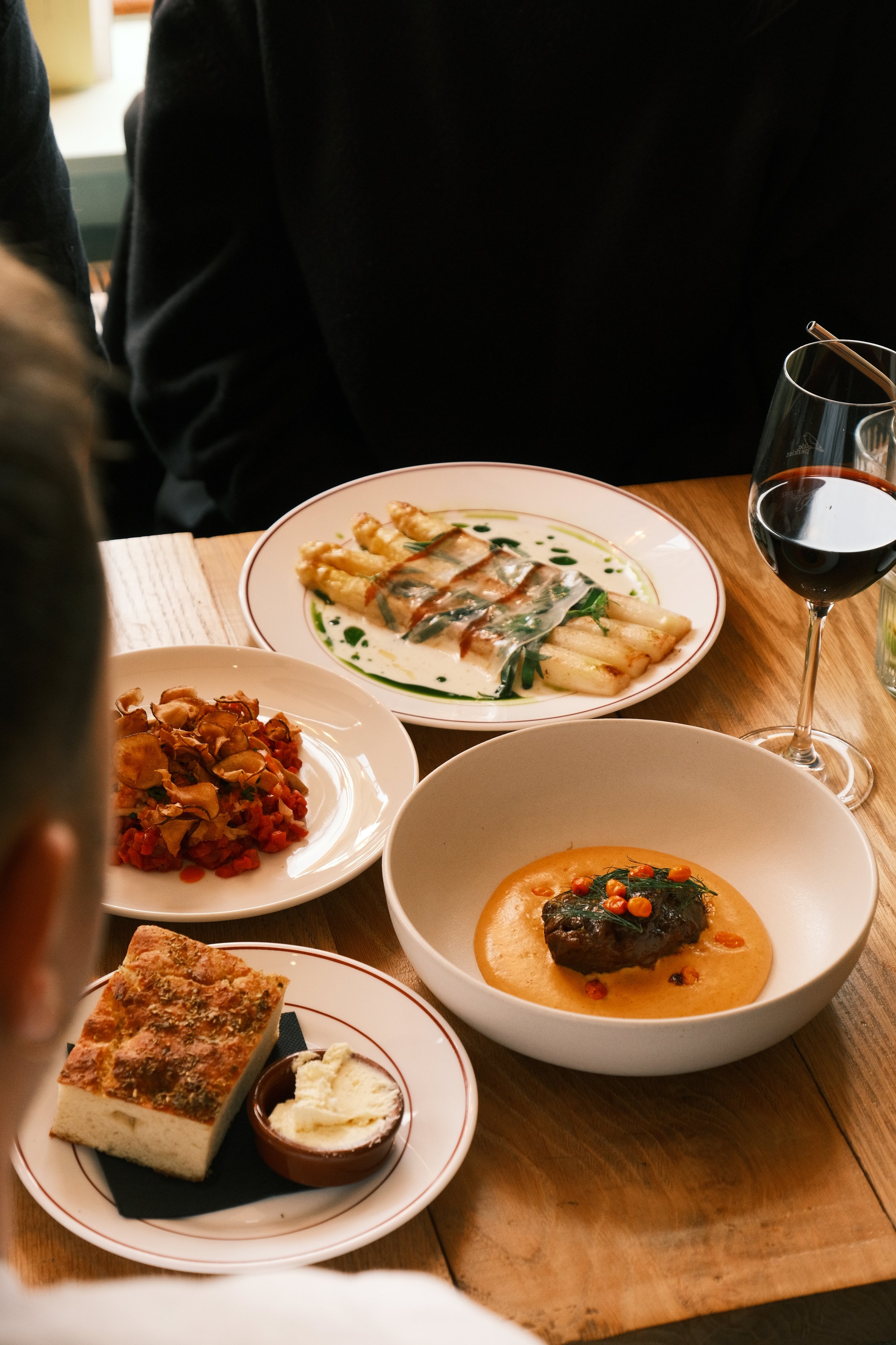 A table with various dishes including pasta with sauce, a bowl of soup with a meat patty and garnish, a slice of cake with cream, and a glass of red wine.