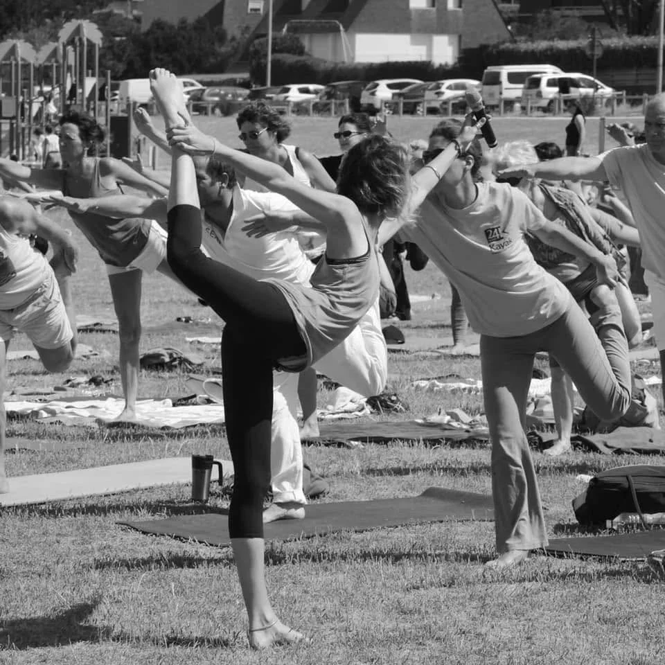 Des personnes pratiquent le yoga en extérieur, effectuant des poses d'équilibre et de flexibilité sur des tapis de yoga, par une journée ensoleillée.
