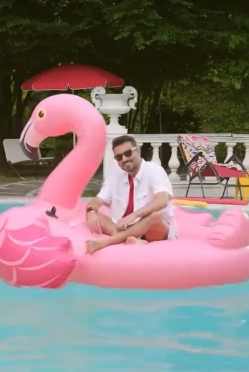 Uomo con occhiali da sole seduto su una grande ciambella gonfiabile a forma di fenicottero rosa nella piscina, con una donna sdraiata sulla ciambella, sullo sfondo una veranda con sedie e un ombrellone rosso.