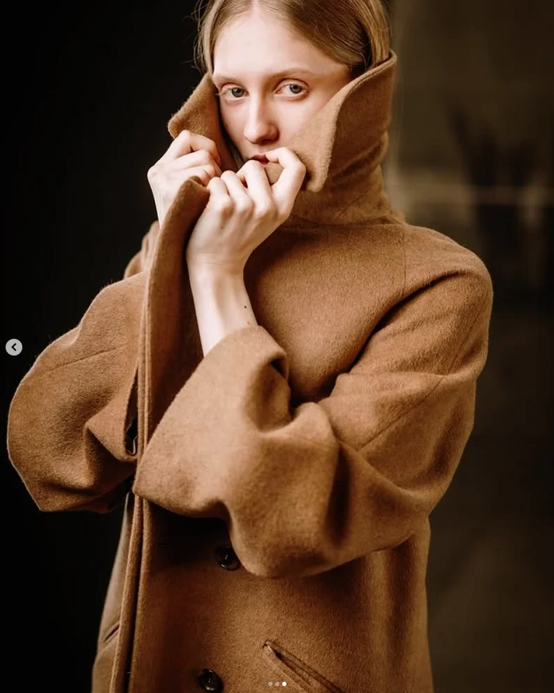 Ragazza con cappotto marrone avvolgente e colletto alzato, che si tiene le mani vicino al volto.