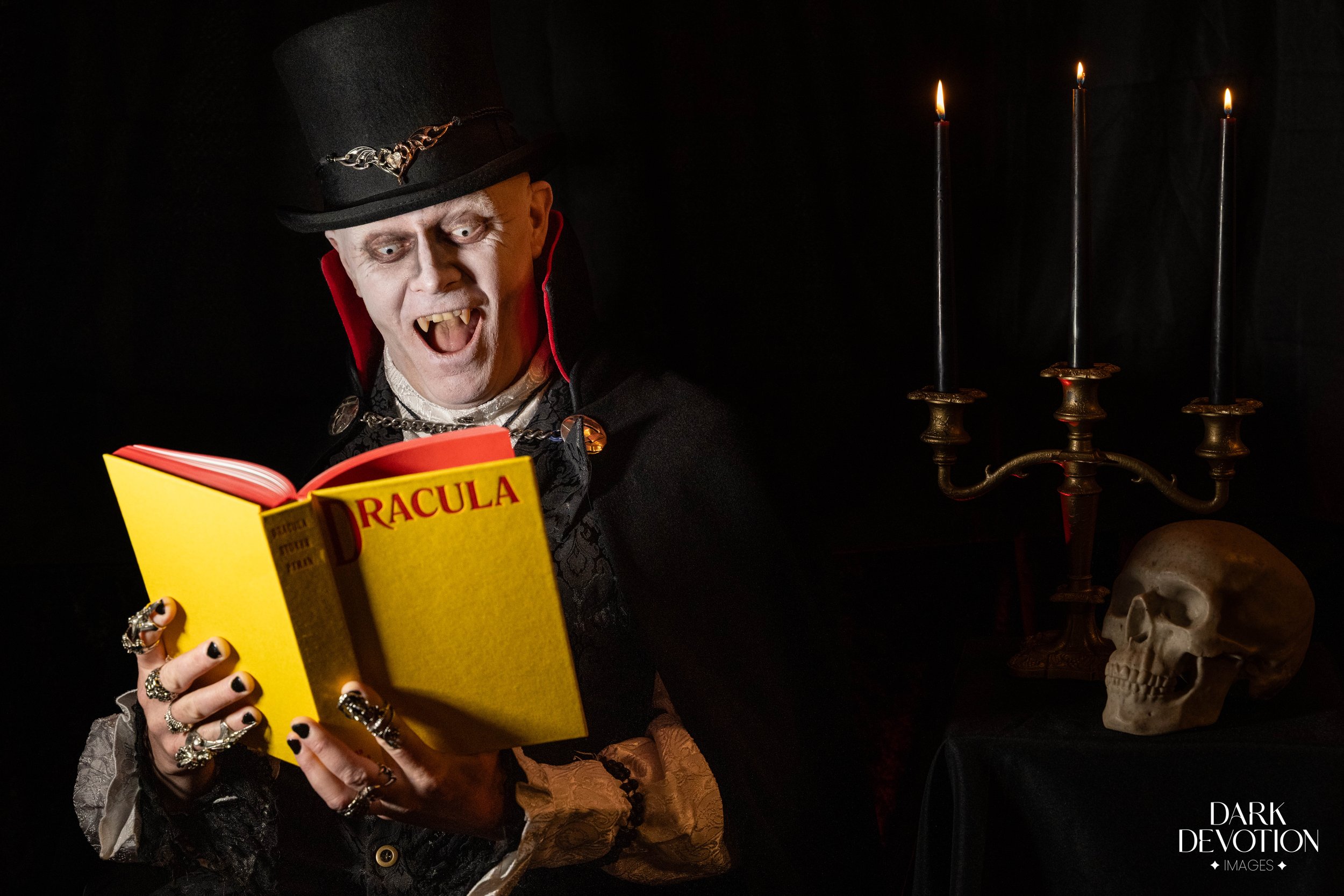 Person dressed as Dracula reading a yellow book titled 'DRACULA' in a dark, gothic setting with candles and skull decorations.