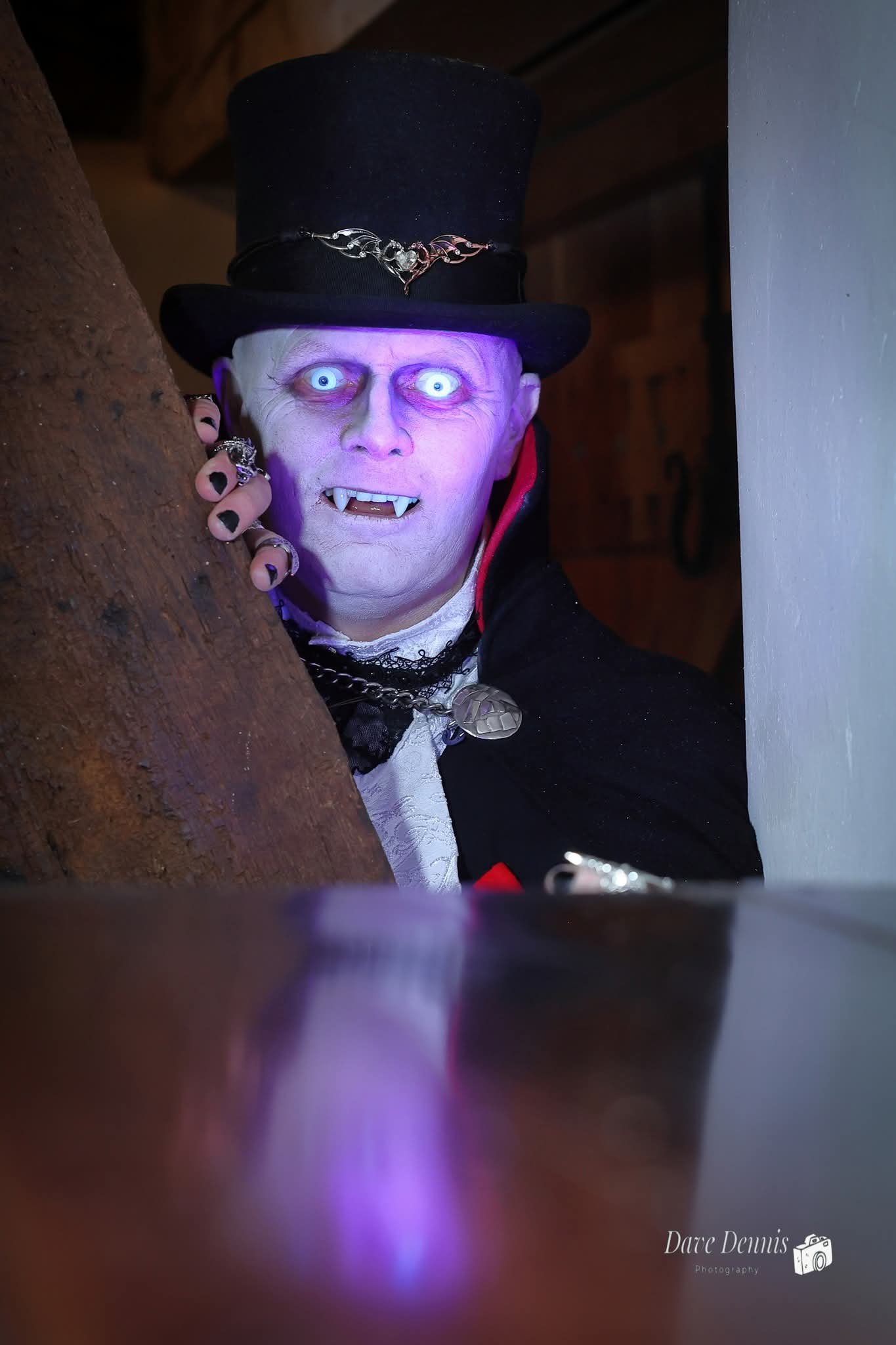 A person dressed as a vampire peeking from behind a wooden surface with a mischievous expression, wearing a black top hat, vampire teeth, and vampire makeup, with hand adorned with rings.