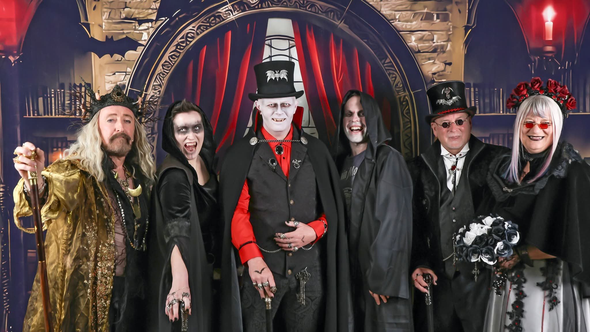 A group of six people dressed in Halloween costumes, standing in front of a spooky backdrop with red curtains and gothic architecture, smiling and posing for the photo.