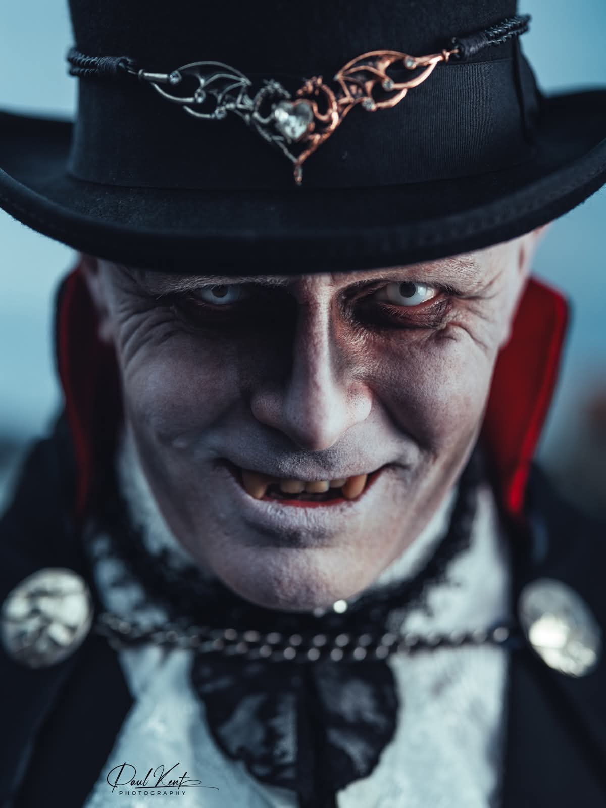 A close-up of a man with pale skin and intense blue eyes, dressed as a vampire with fangs, wearing a black top hat decorated with a metallic chain and skulls, and a black outfit with a white shirt and black bow tie, snarling with a menacing expression.