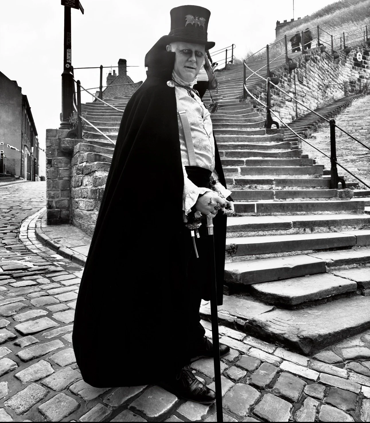 A person dressed as a gothic vampire with pale face makeup, wearing a tall top hat with bat designs, a long black cape, ornate rings, and holding a cane, standing on a cobblestone street near a staircase in black and white photograph.