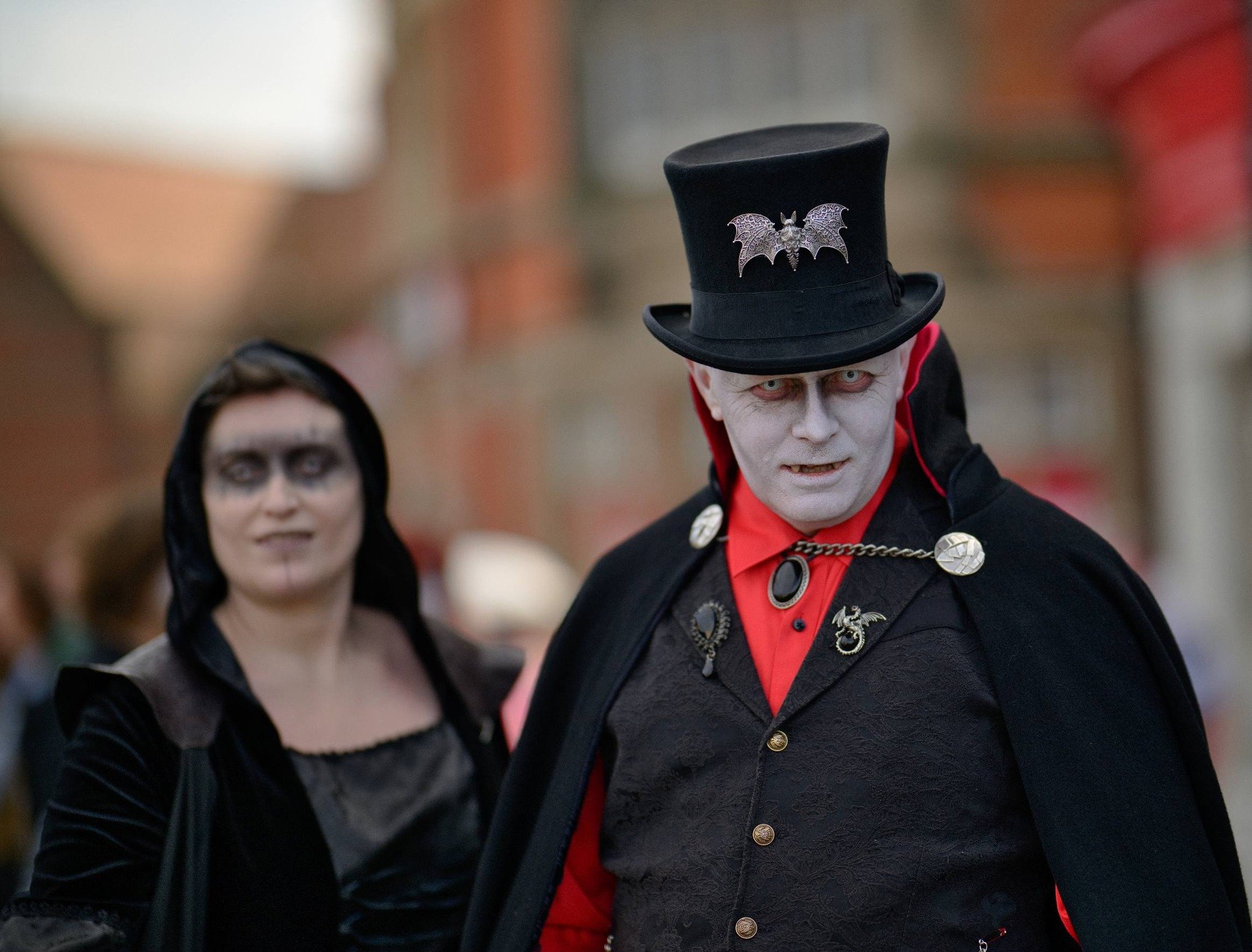 Costumes of two individuals dressed as vampires or Gothic characters, with pale faces, dark clothing, and gothic accessories, in a festive outdoor setting.