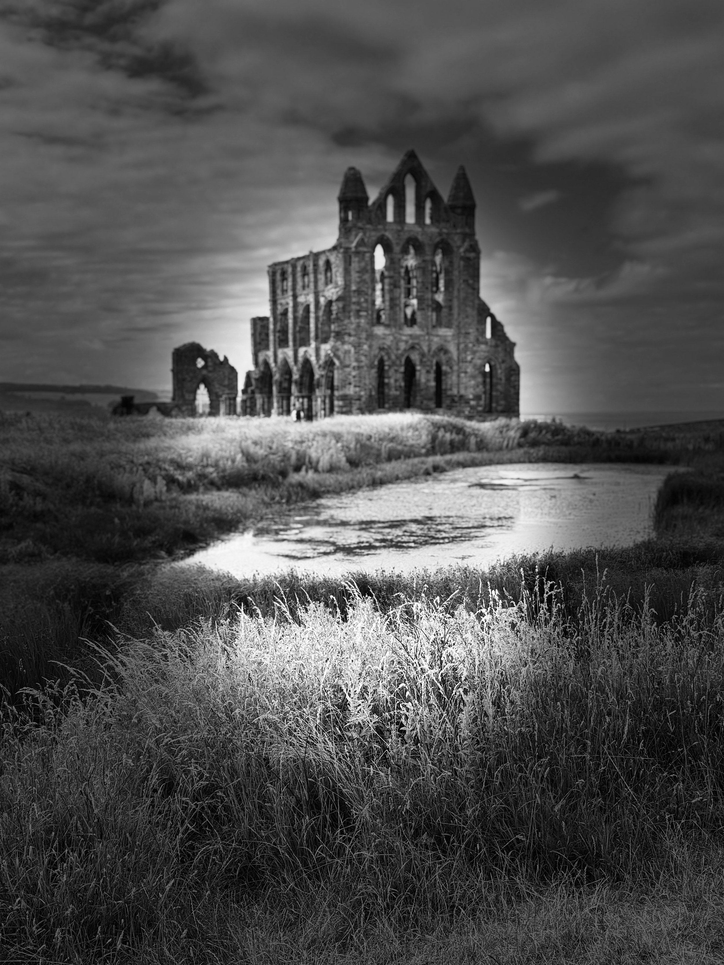 Black-and-white photograph of a Gothic stone abbey ruin beside a stream under stormy clouds