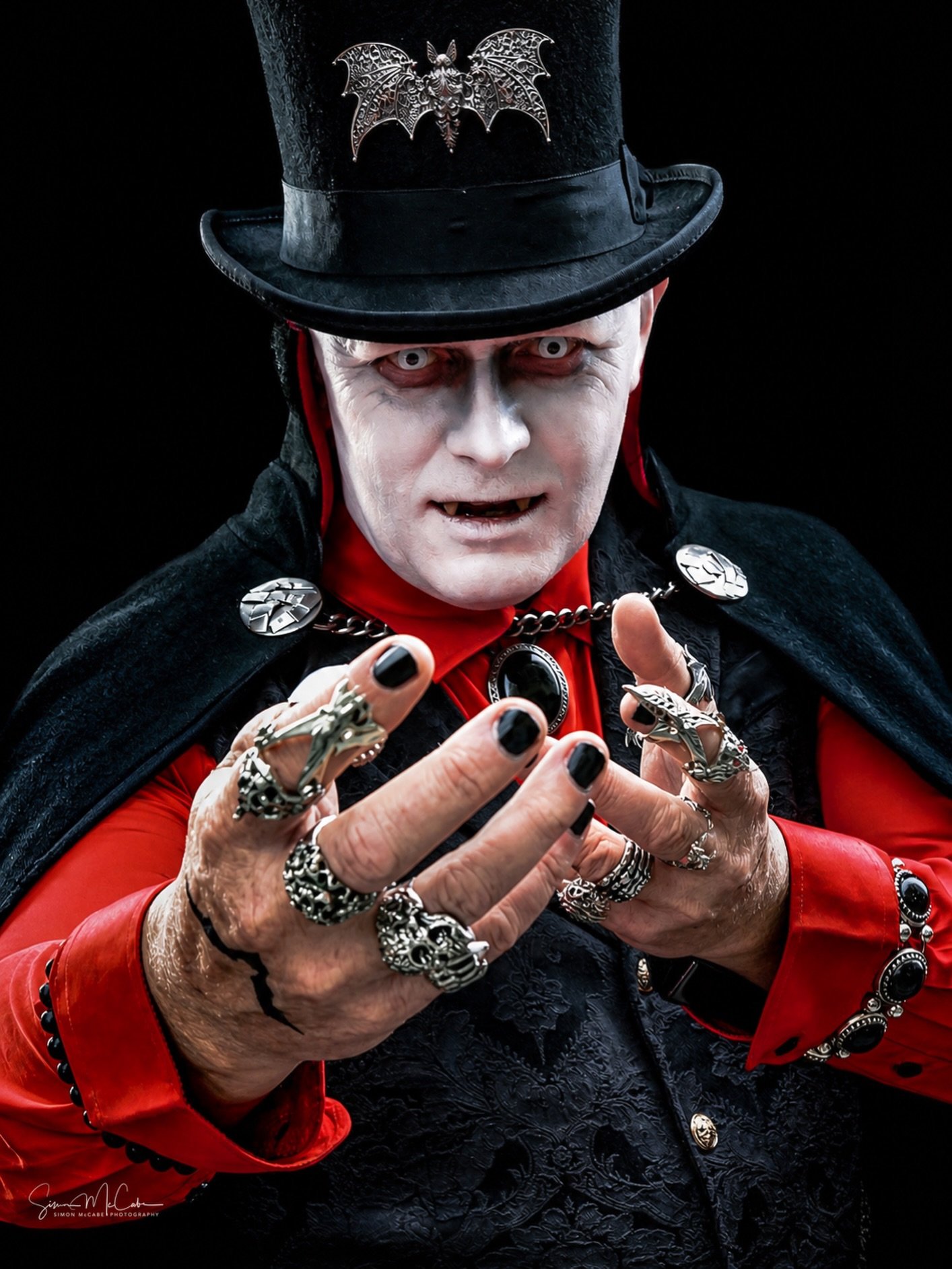 A person dressed as a vampire with a black top hat decorated with a metal bat, wearing white face makeup, red contact lenses, and black lipstick. They are wearing a red shirt, black jacket, and a black cape with silver brooches, and have multiple silver rings and accessories on their hands.
